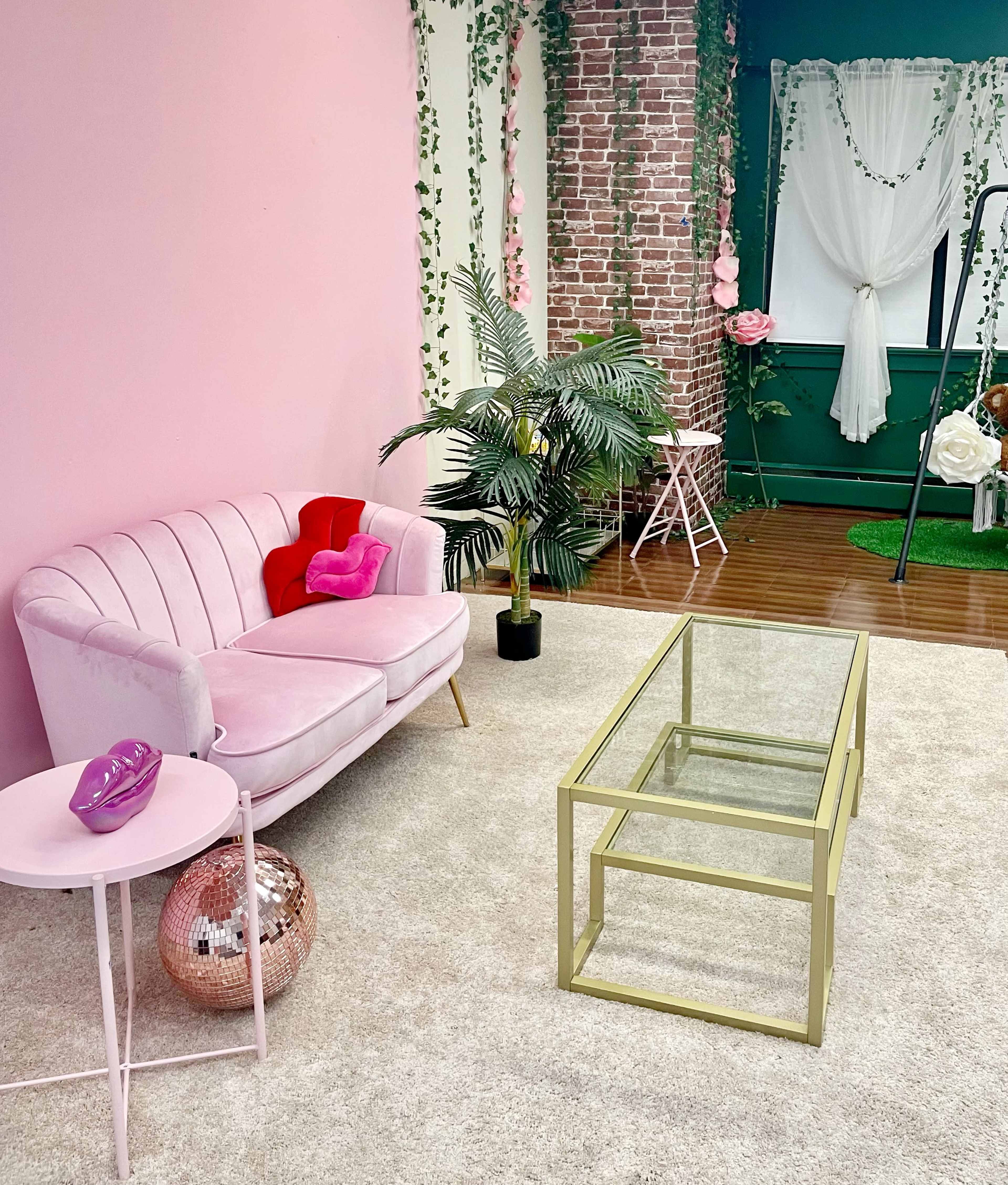 A pink sofa, a glass top table, and a potted plant in a decorated room with a pink wall and a brick accent wall.