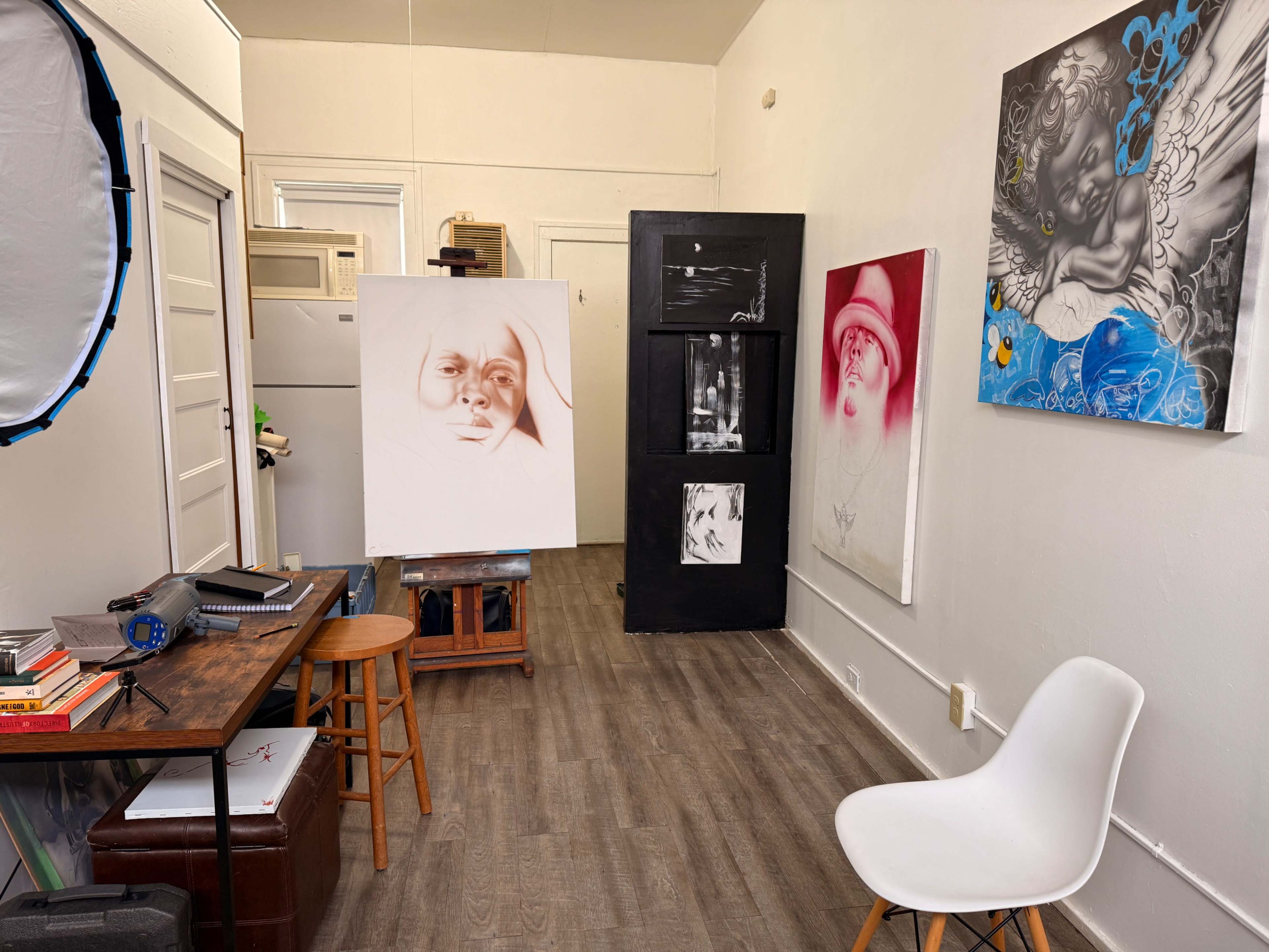 The image shows an artist's studio with unfinished canvases, a black storage unit, and paintings displayed on the walls.