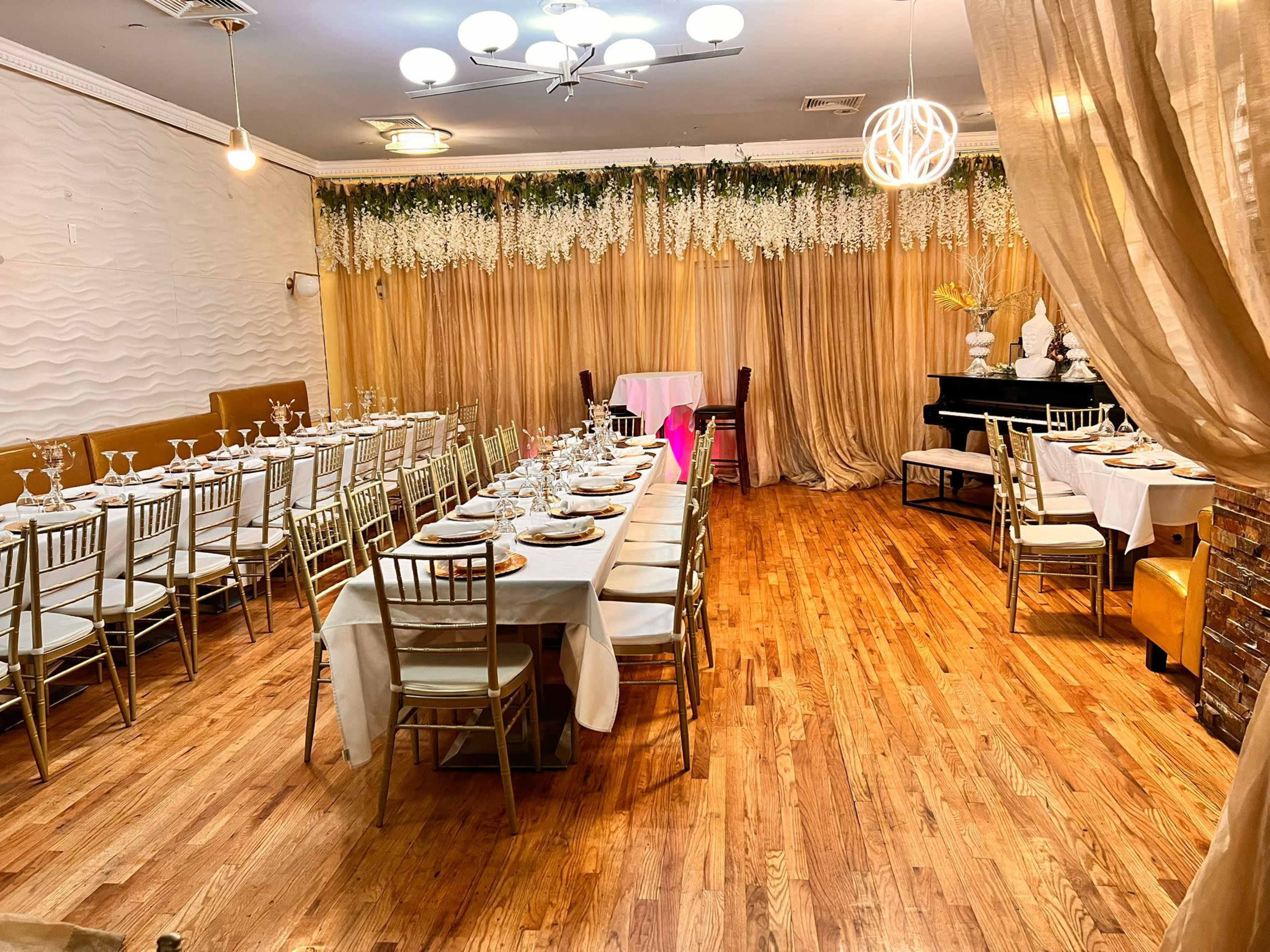 The image shows a decorated dining area with two long tables set for a formal event, featuring golden chairs and elegant tableware under soft lighting and draped fabrics.