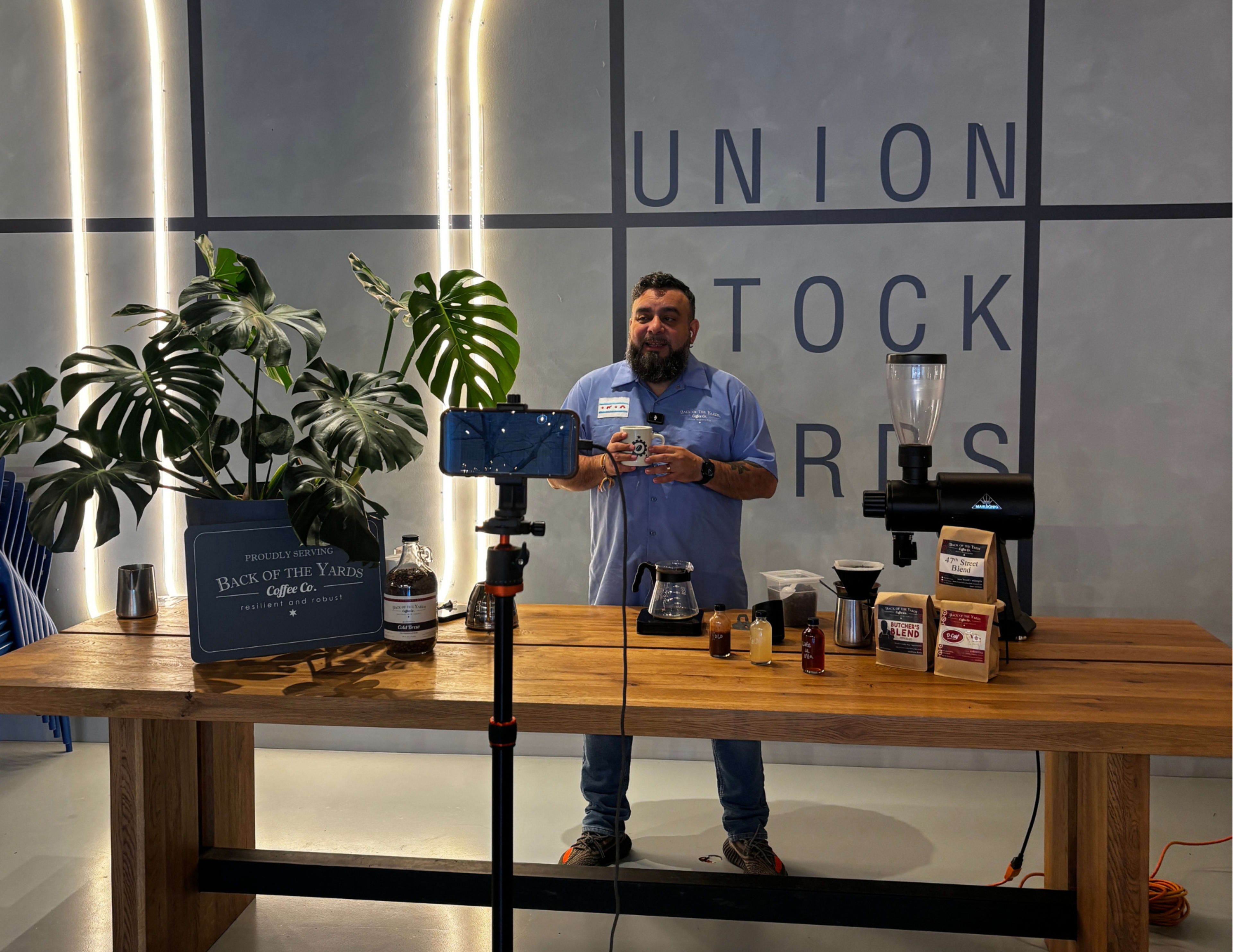 A man stands behind a wooden table with various coffee-making tools and ingredients, speaking into a camera set on a tripod in a space labeled "Union Stock Yards."