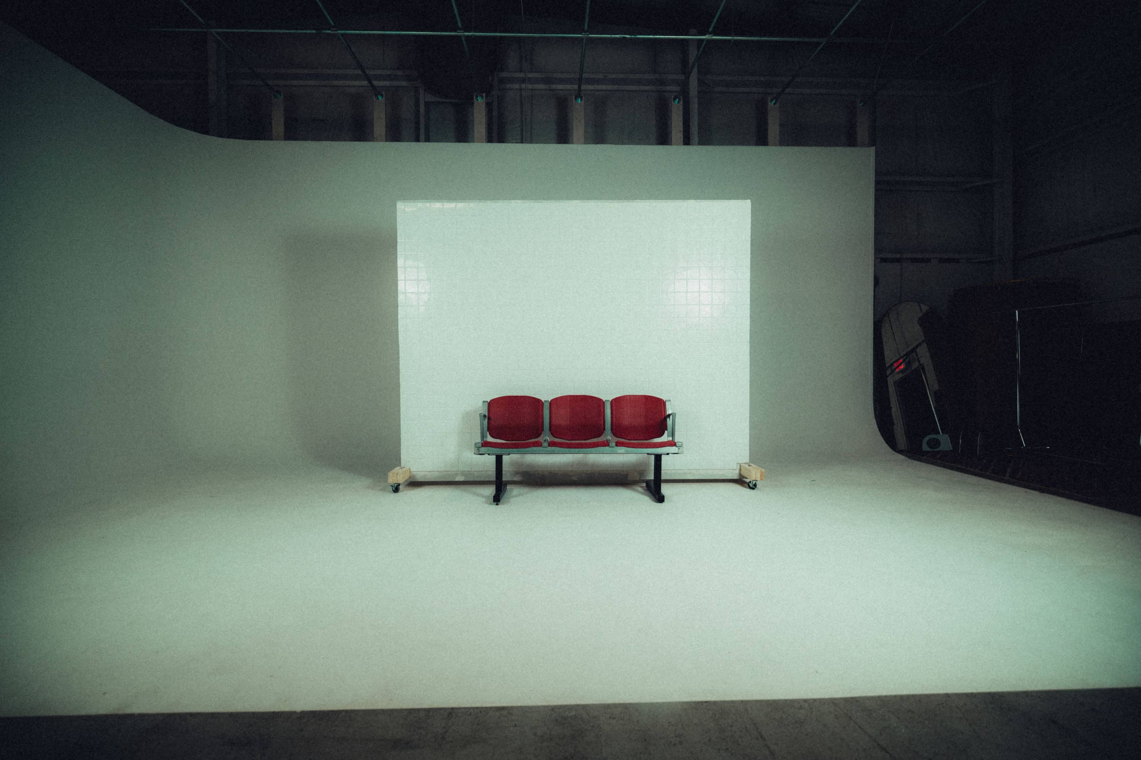 A row of three red chairs is positioned in front of a large white backdrop in a spacious studio environment.