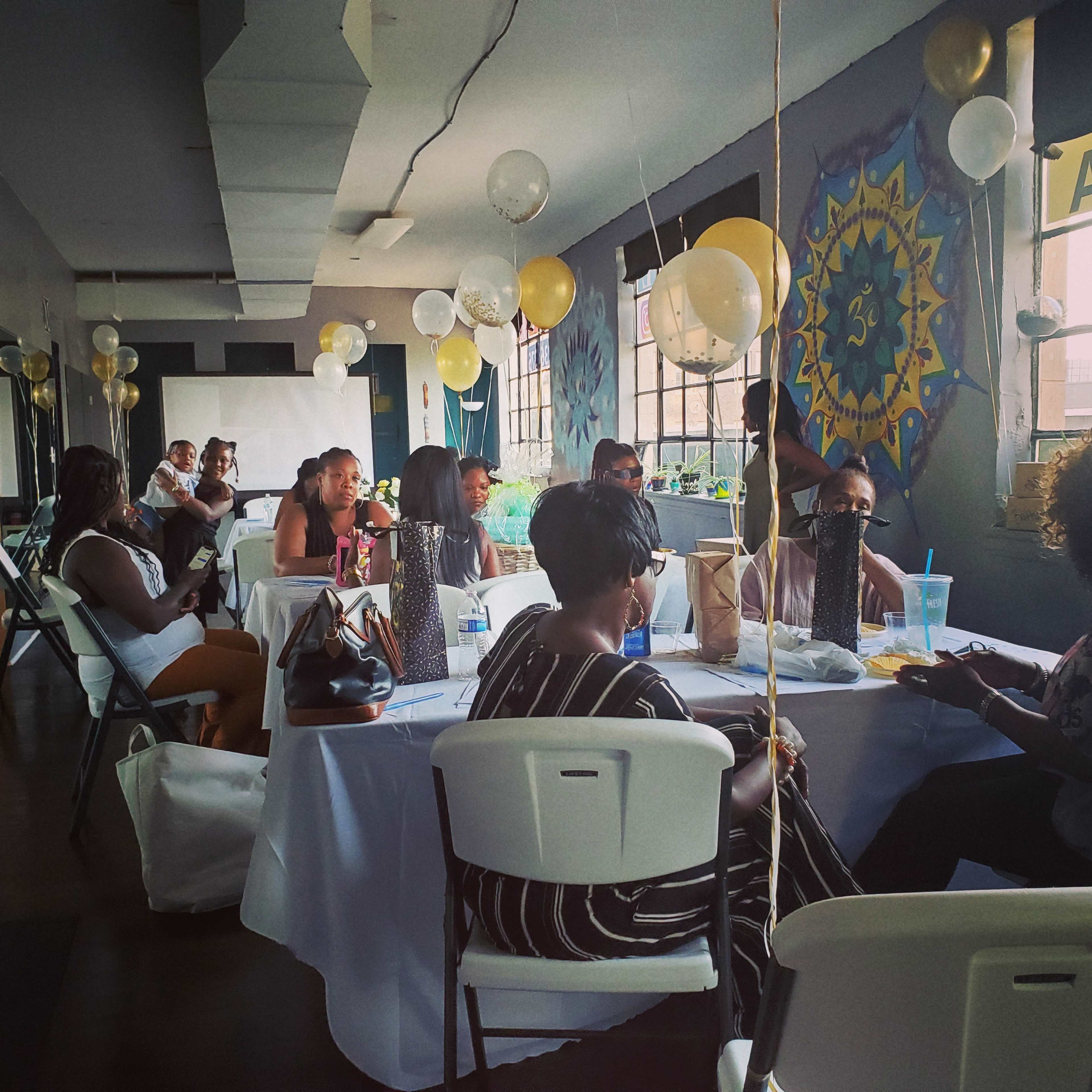 A group of people sit around tables in a decorated room with balloons and wall art, engaged in conversation.