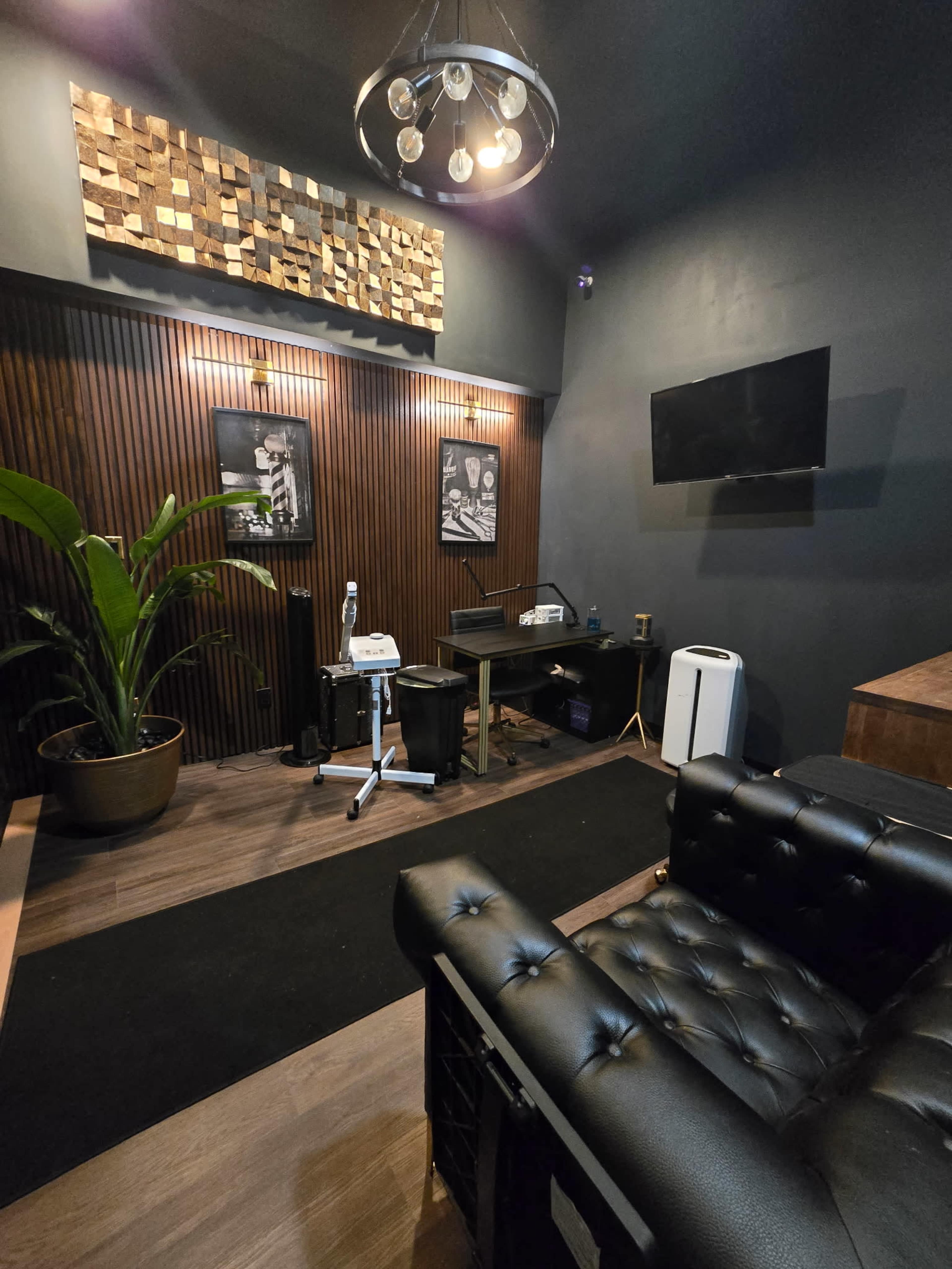 The image shows a modern, minimalist office space with a black leather couch, a desk with office equipment, a large plant, and artwork mounted on the walls.