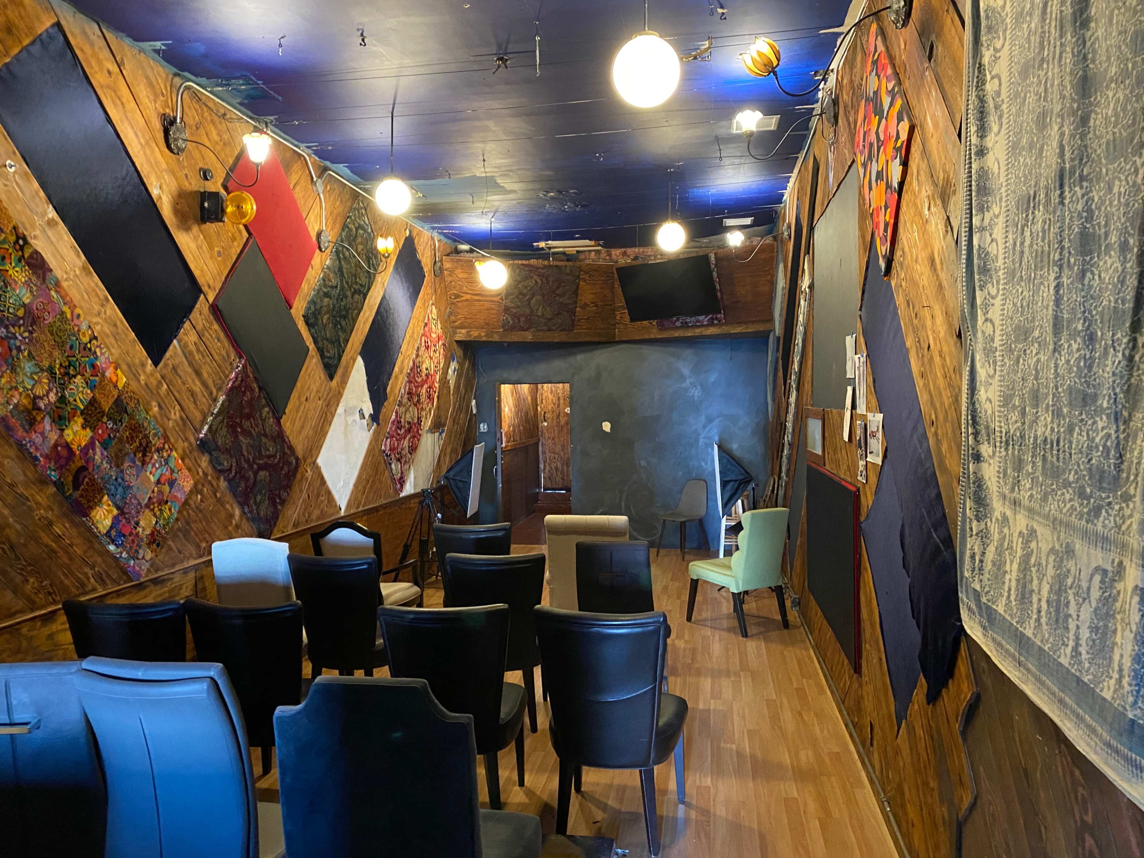 A sparsely furnished room with wooden walls adorned with colorful fabric panels, several chairs arranged in rows, and various lights hanging from the ceiling.