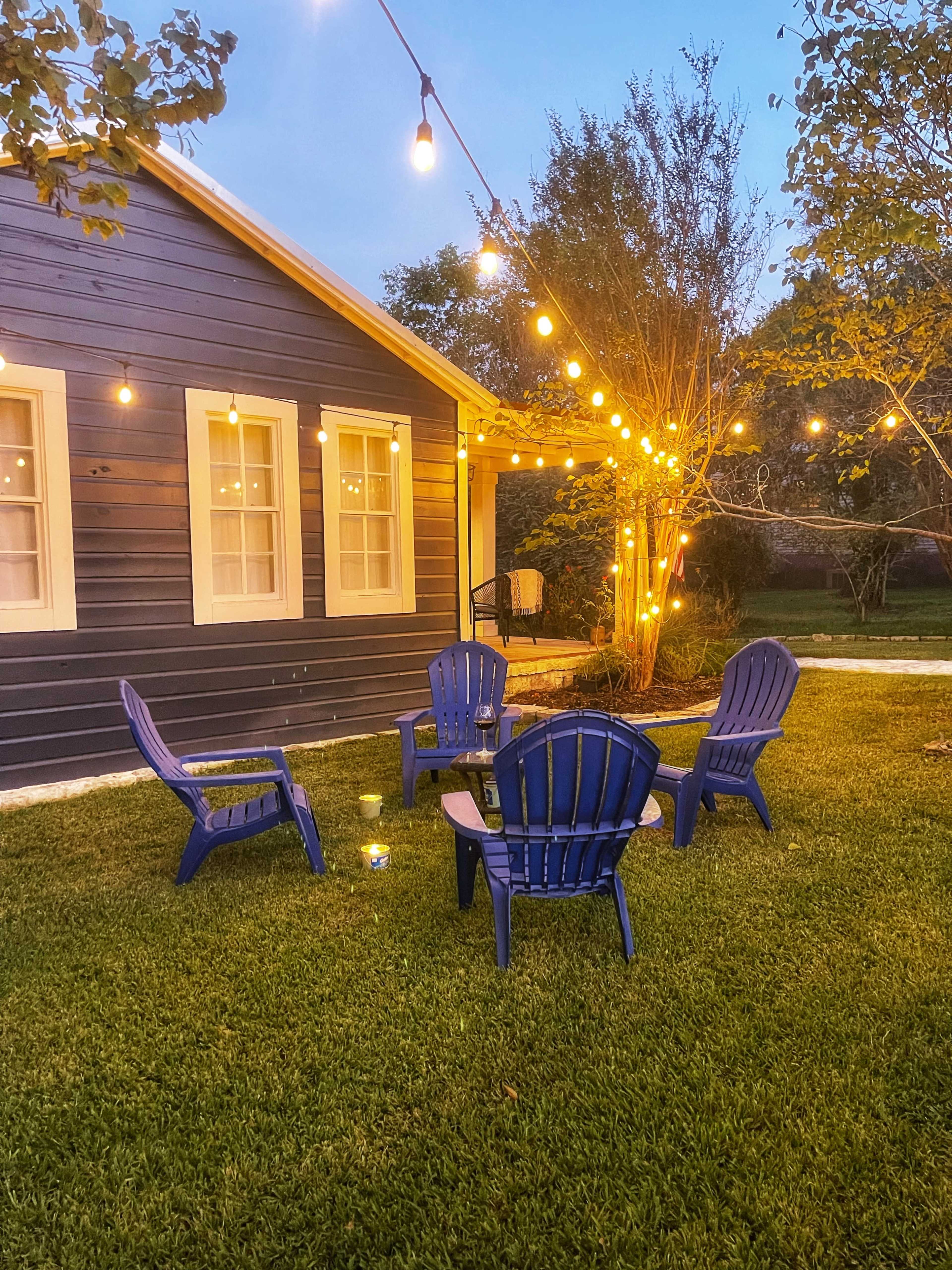 A cozy outdoor seating area features four blue Adirondack chairs arranged in a circle under string lights near a blue house.