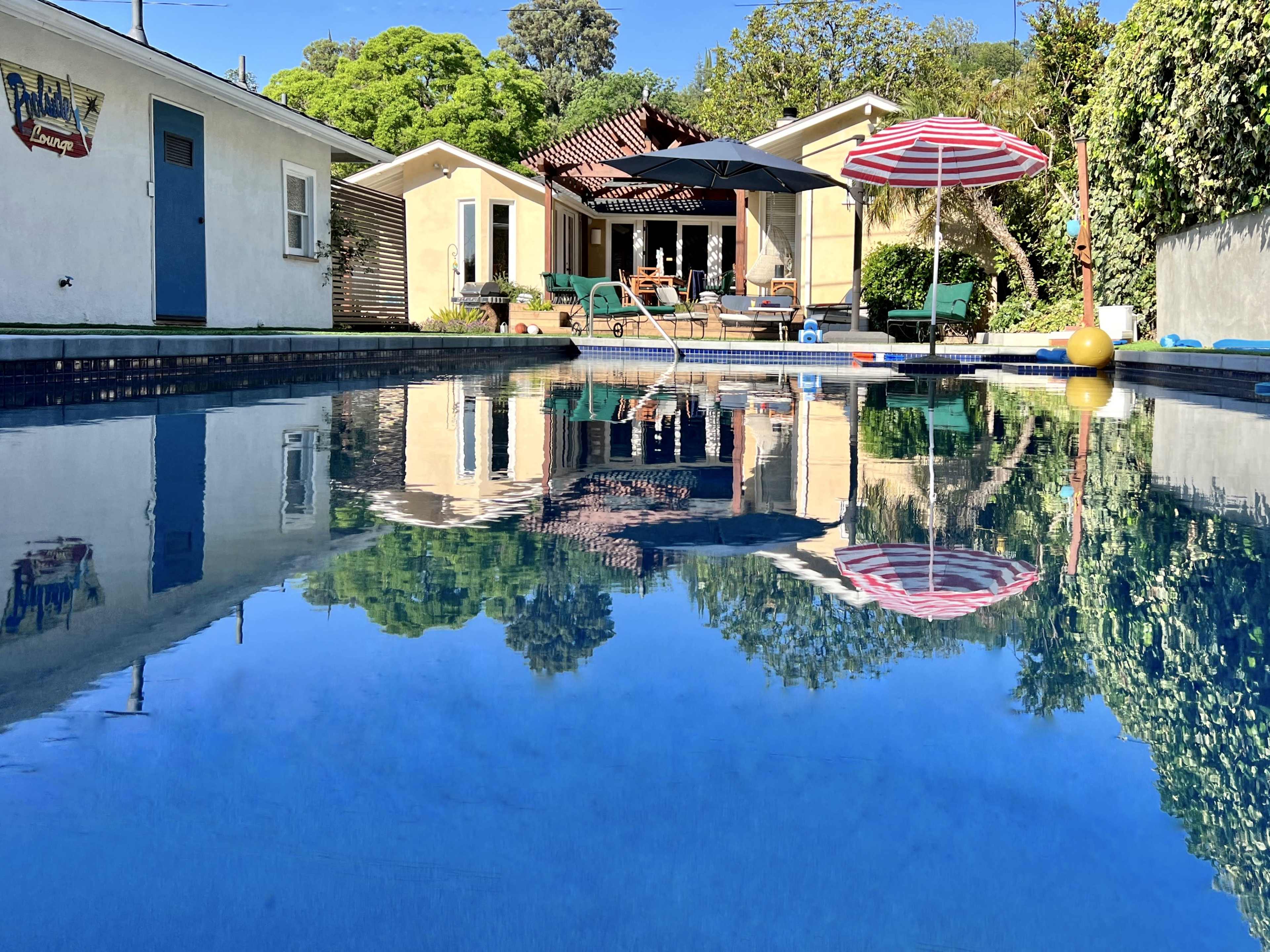 A clear blue pool reflects a sunny backyard with a house, patio, and outdoor furniture surrounded by trees.