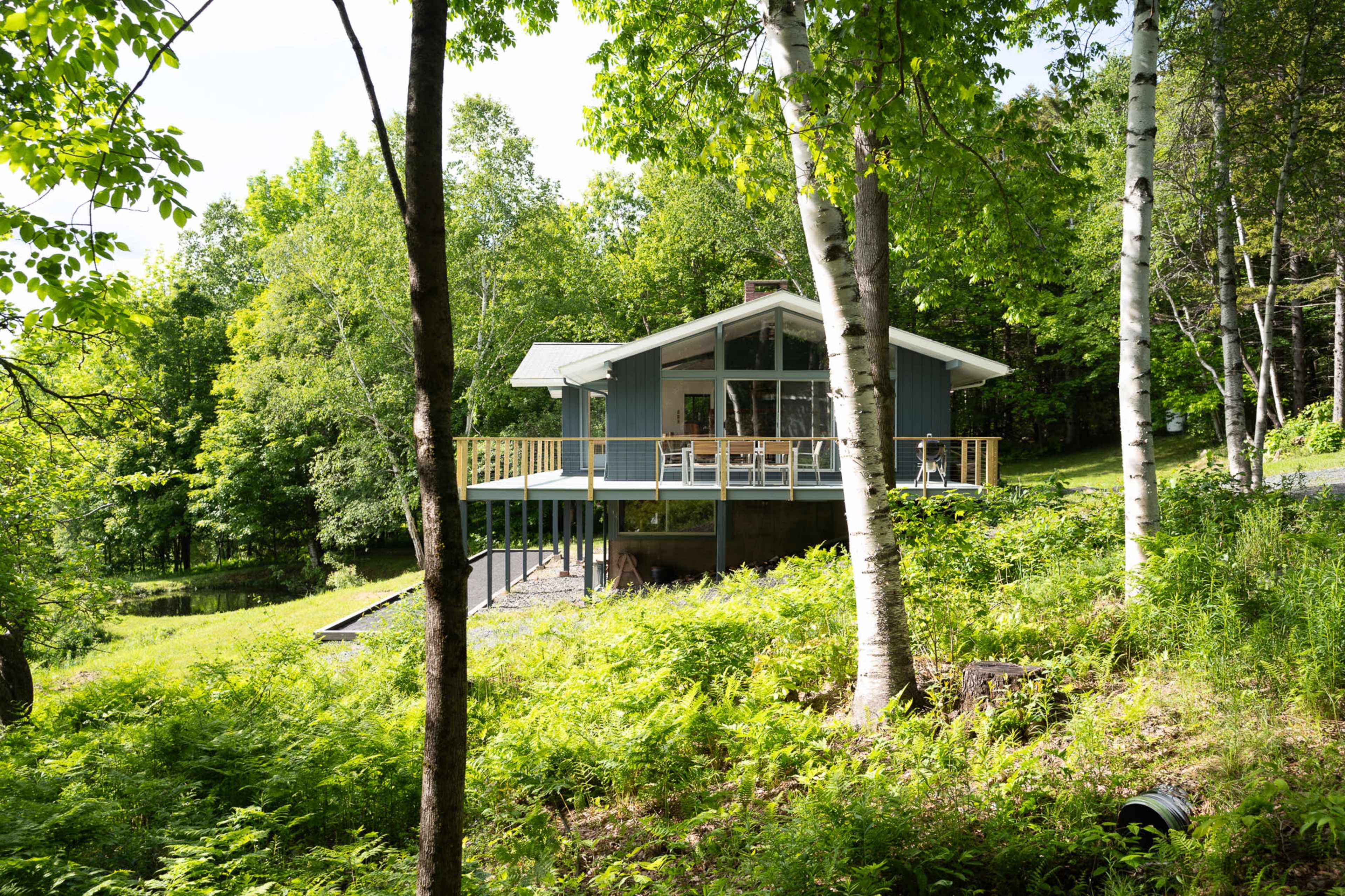 A modern house with large windows and a deck is situated on a grassy hillside surrounded by trees.