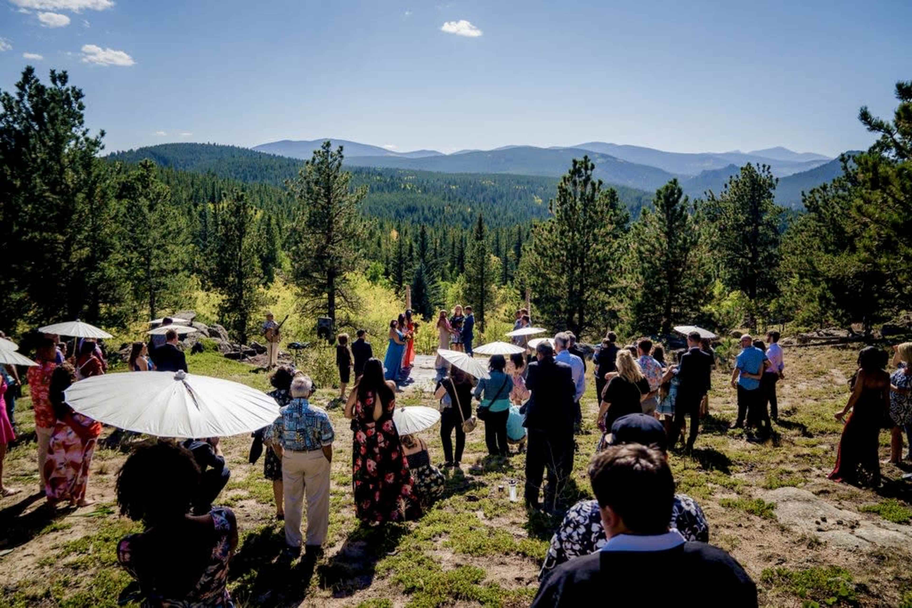 A wedding ceremony takes place outdoors in a mountainous area, surrounded by guests holding umbrellas and trees.
