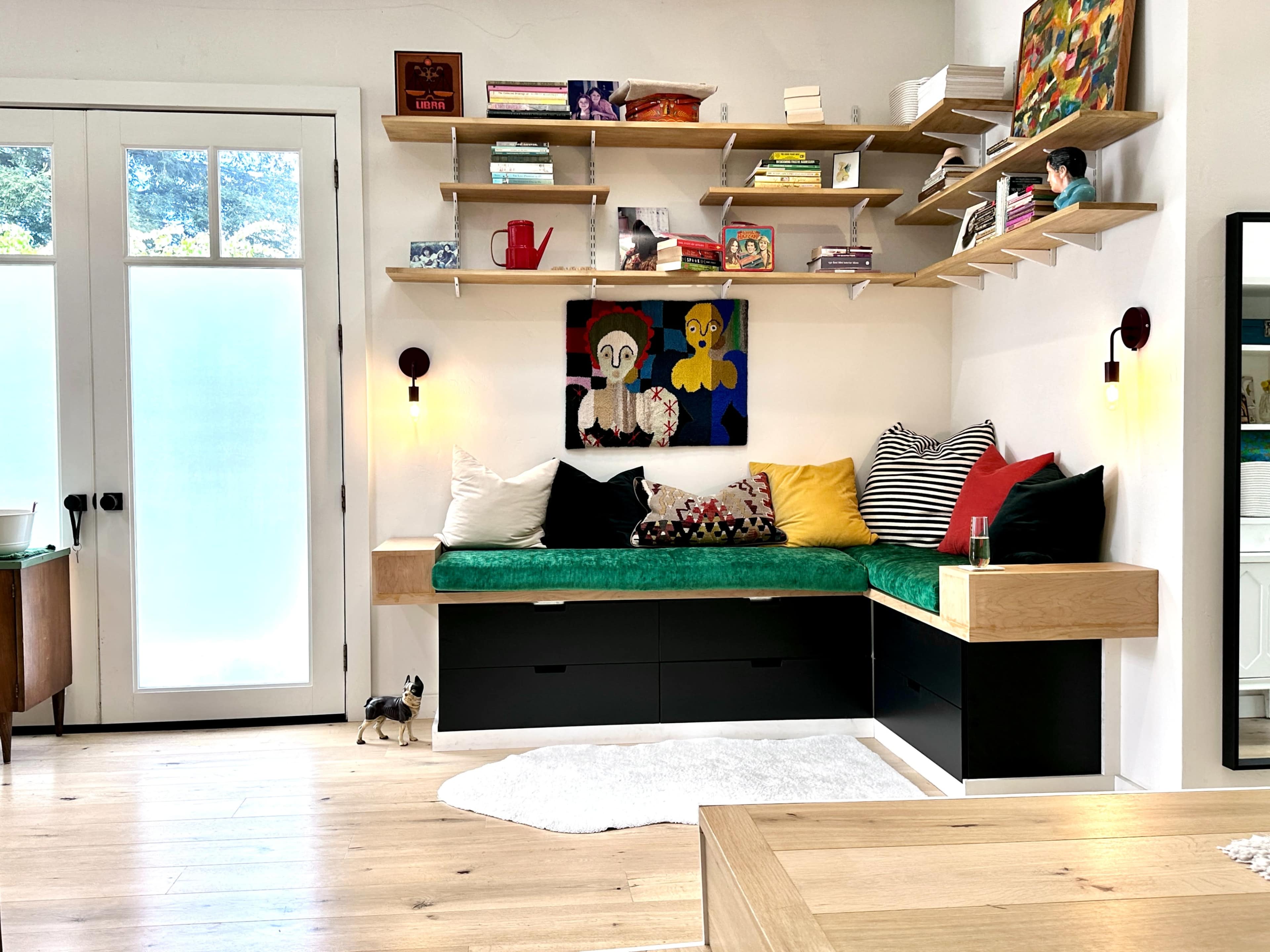 A modern room featuring a green cushioned window seat, shelves with various items and artwork, and light-colored wooden flooring.
