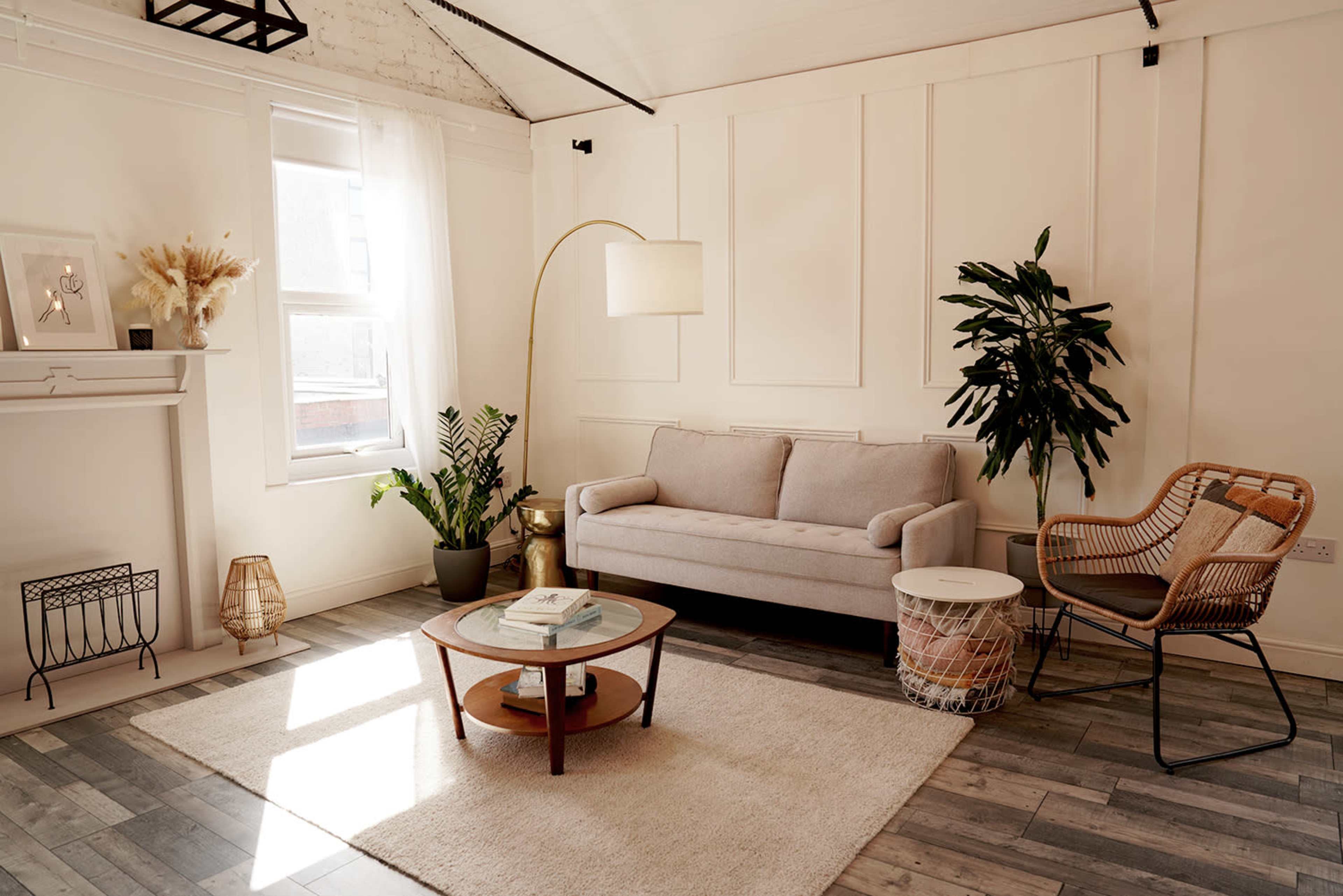 The image shows a modern living room featuring a light-colored sofa, a round coffee table, a floor lamp, and a large potted plant, set against a backdrop of white walls and wooden flooring.