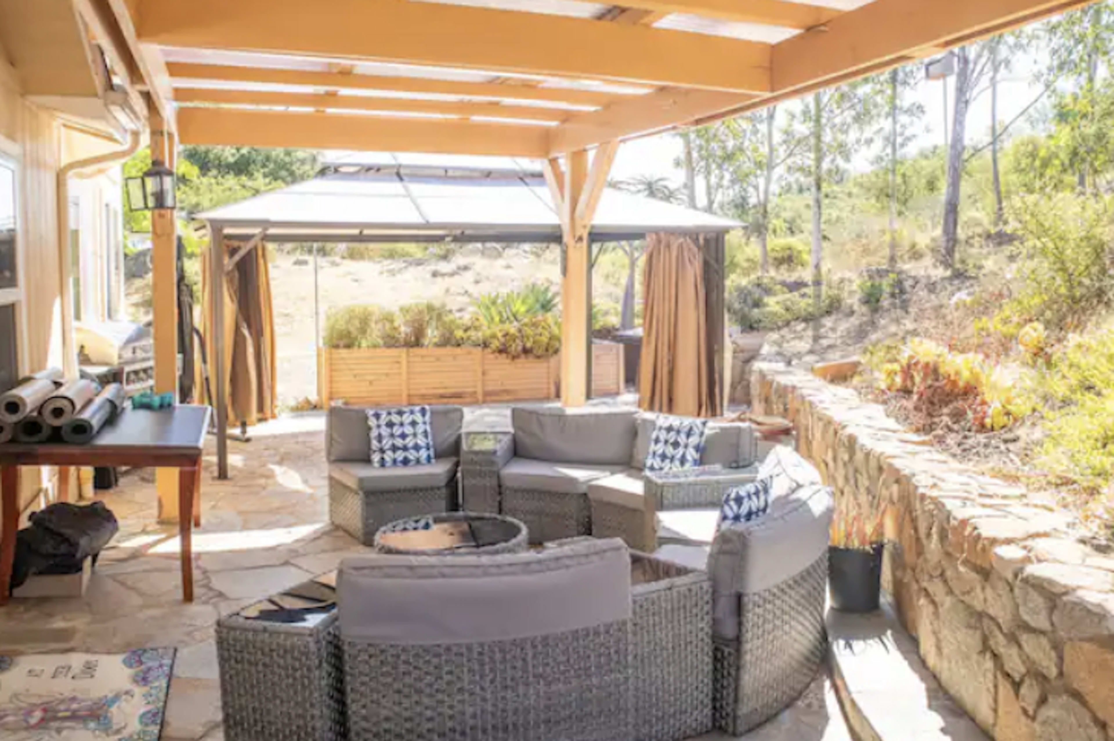 The image shows a shaded outdoor seating area with wicker sofas arranged around a circular table, adjacent to a gazebo and surrounded by greenery.