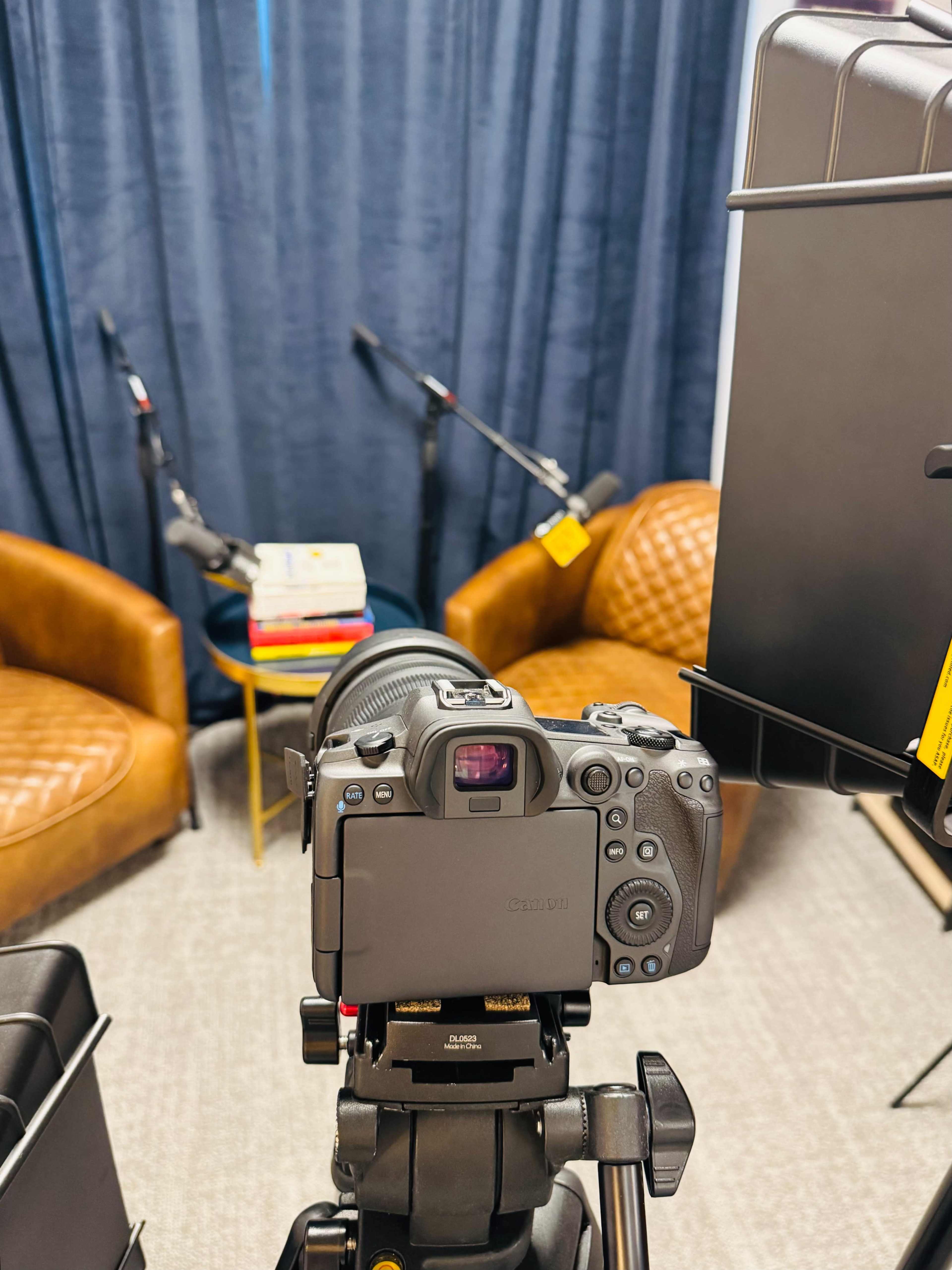 The image shows a camera set up on a tripod facing two brown armchairs and a small table, with microphones positioned nearby in a studio-like setting.