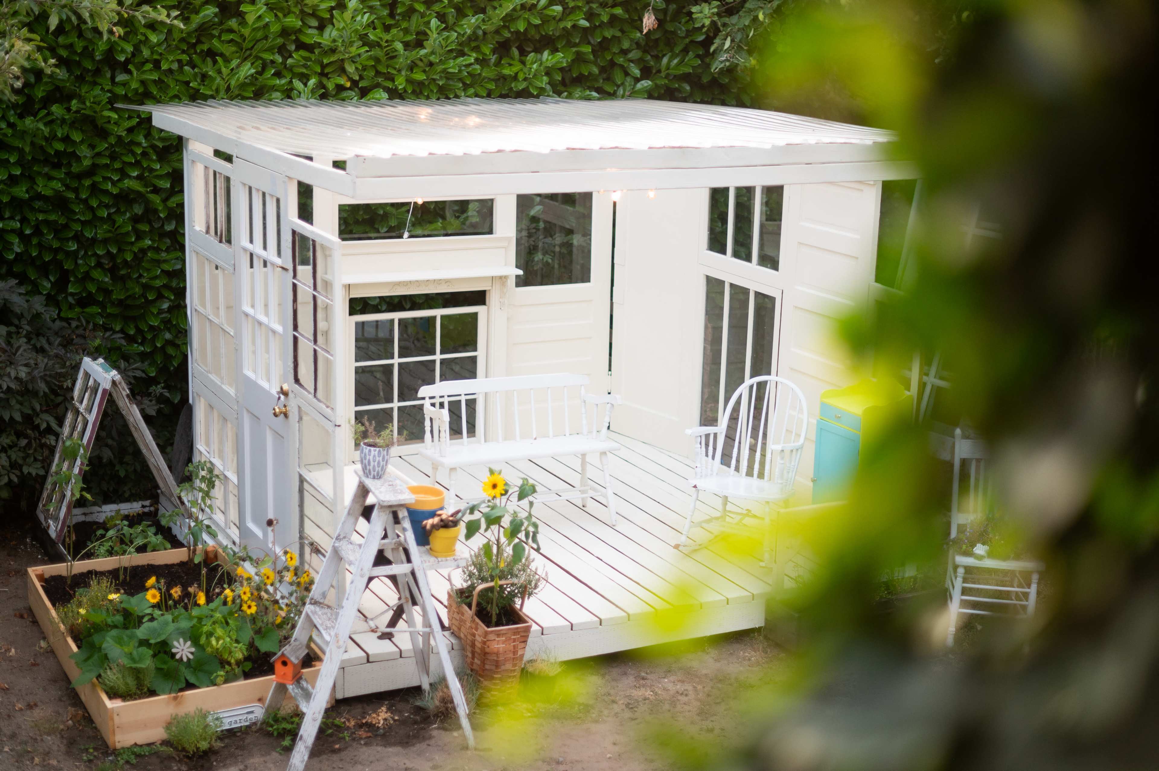 A small white shed with large windows is surrounded by a flower garden and a ladder, with a chair and potted plants on a wooden deck.