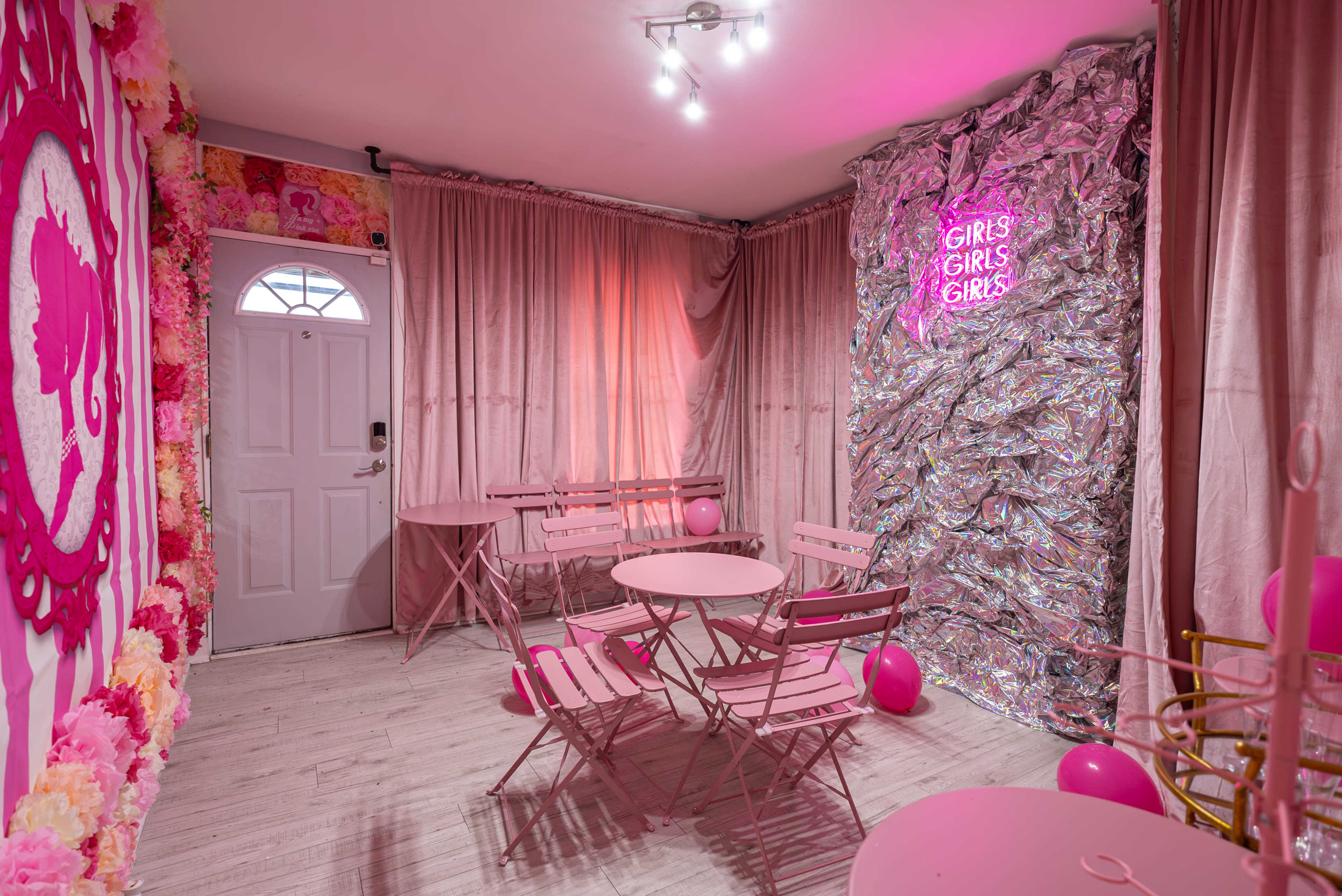 The room features pink décor with folding tables and chairs, a neon sign that reads "GIRLS GIRLS GIRLS," and floral accents.