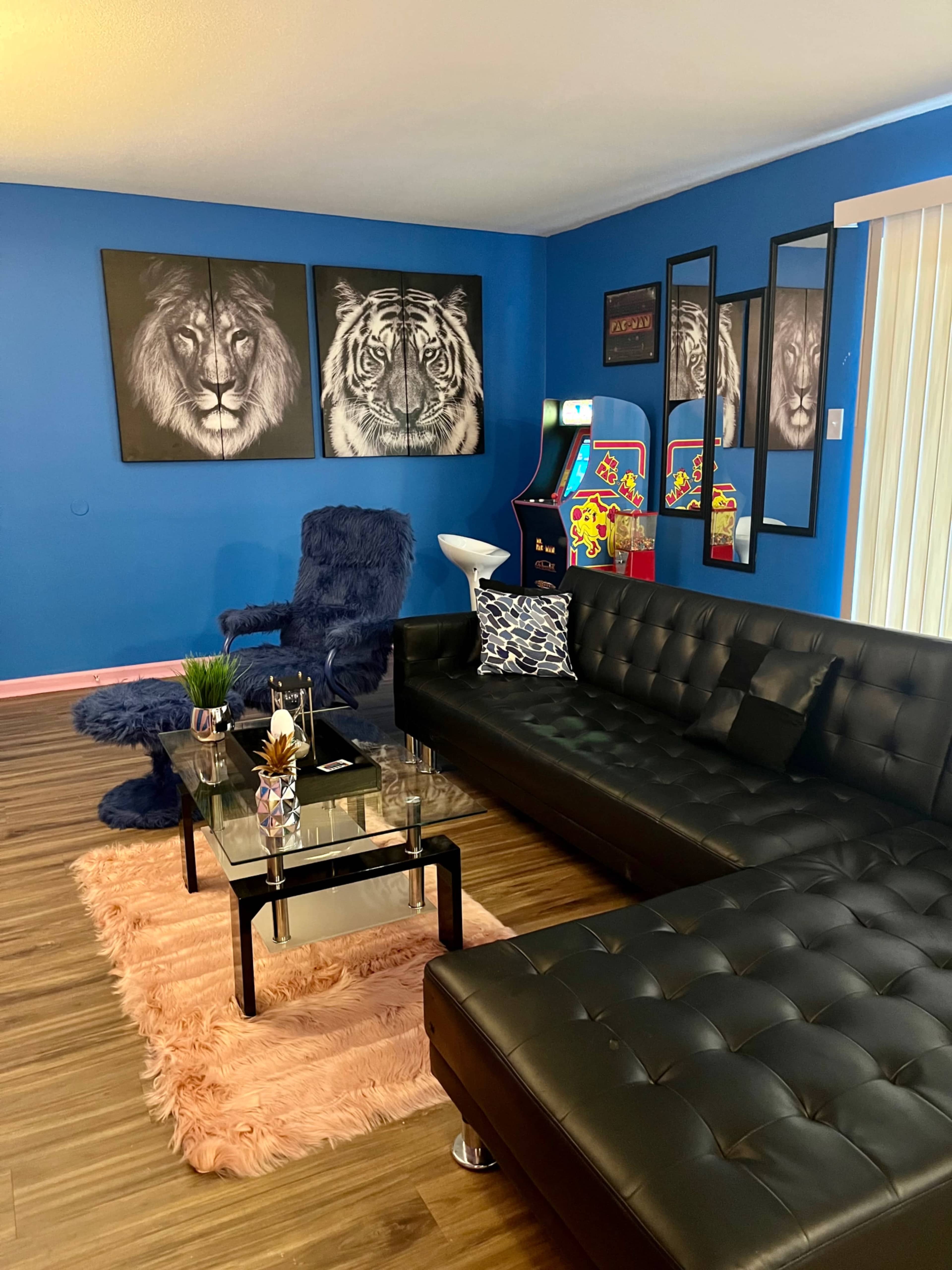 The living room features a black leather sectional sofa, a glass coffee table, and vibrant blue walls adorned with tiger artwork, alongside a vintage arcade game and a plush chair.