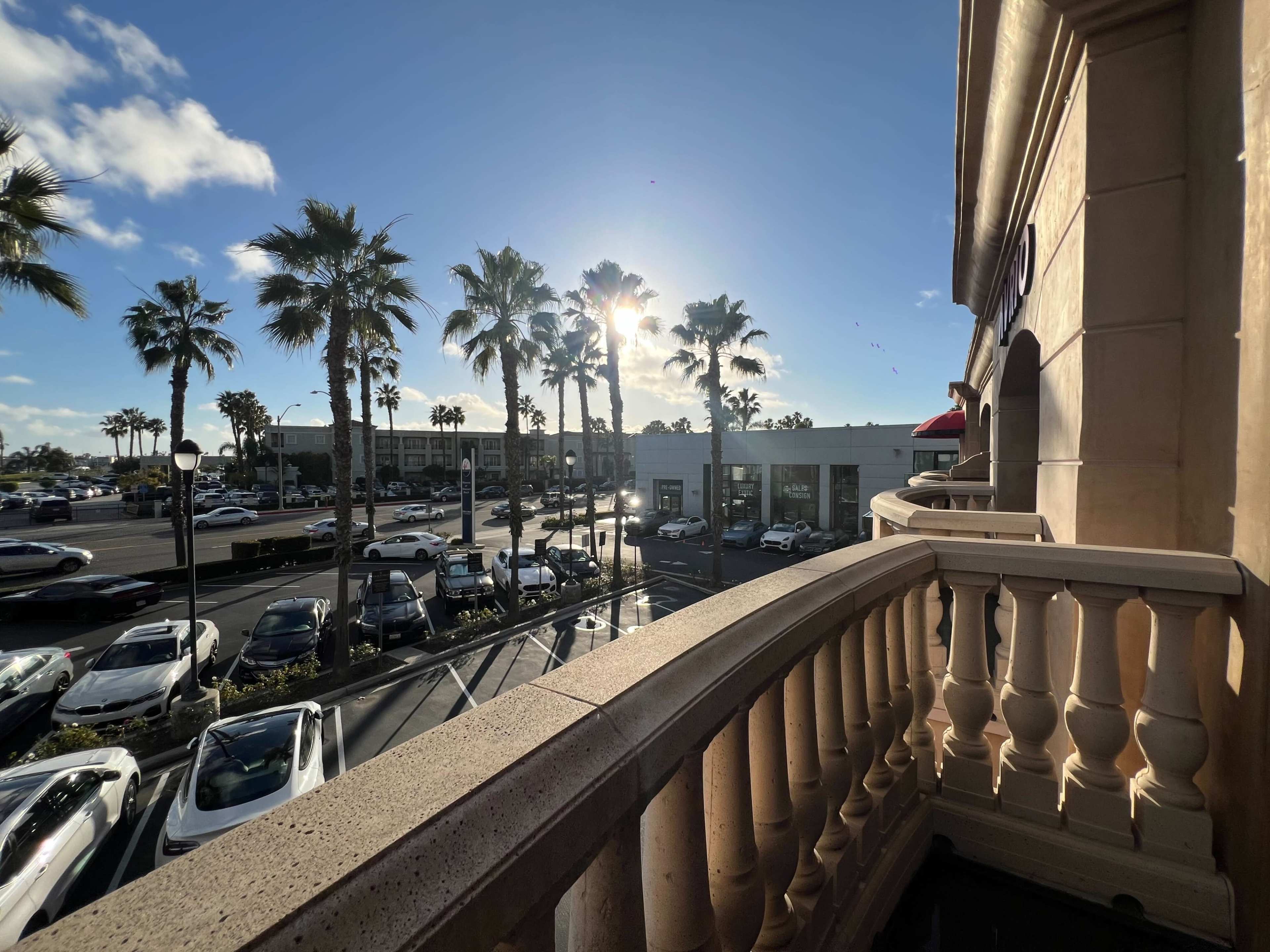 A view from a balcony overlooking a parking lot lined with palm trees and buildings under a clear sky with sunlight.