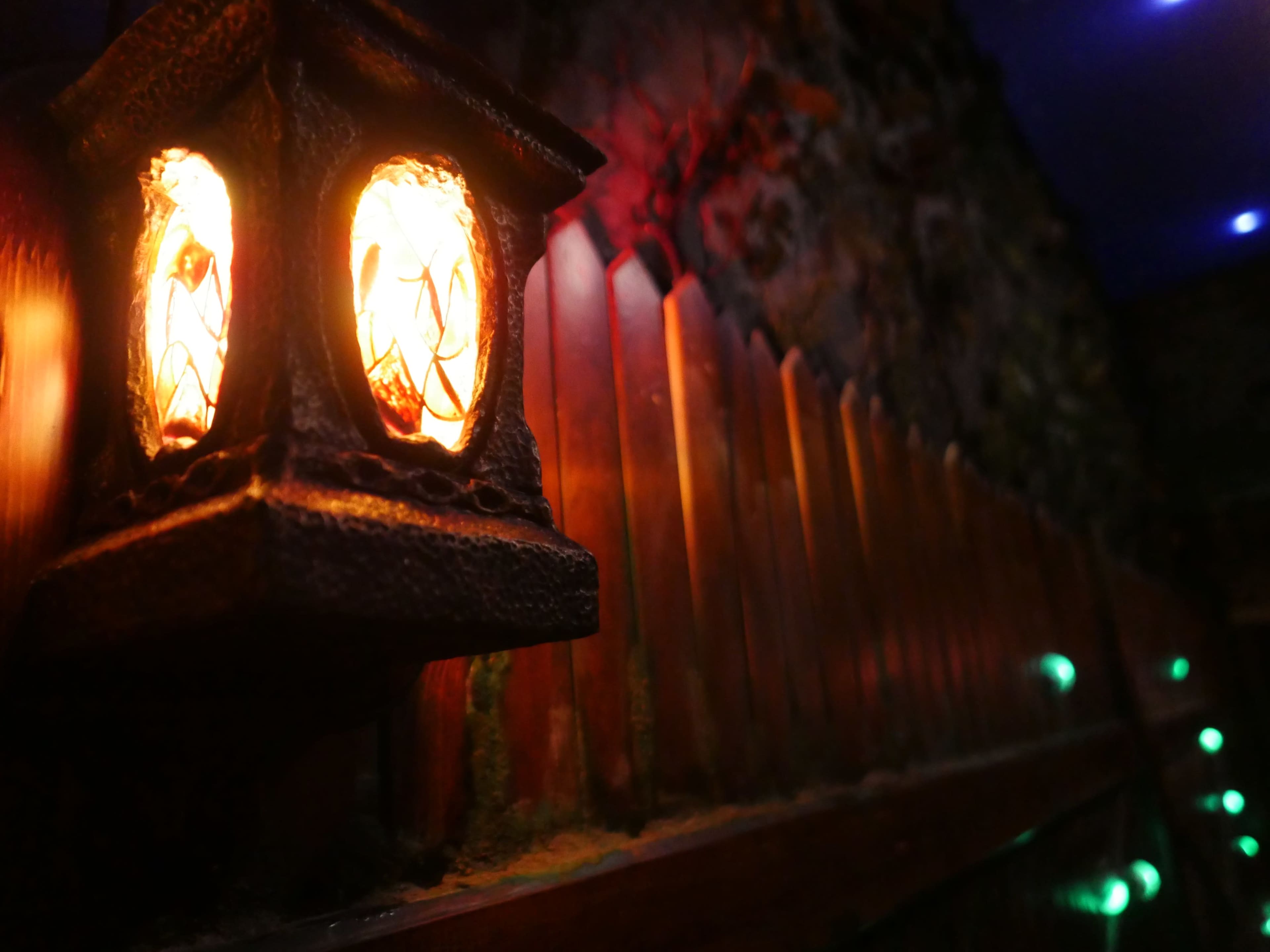 A lantern with two glowing bulbs is mounted next to a wooden fence in a dimly lit setting.