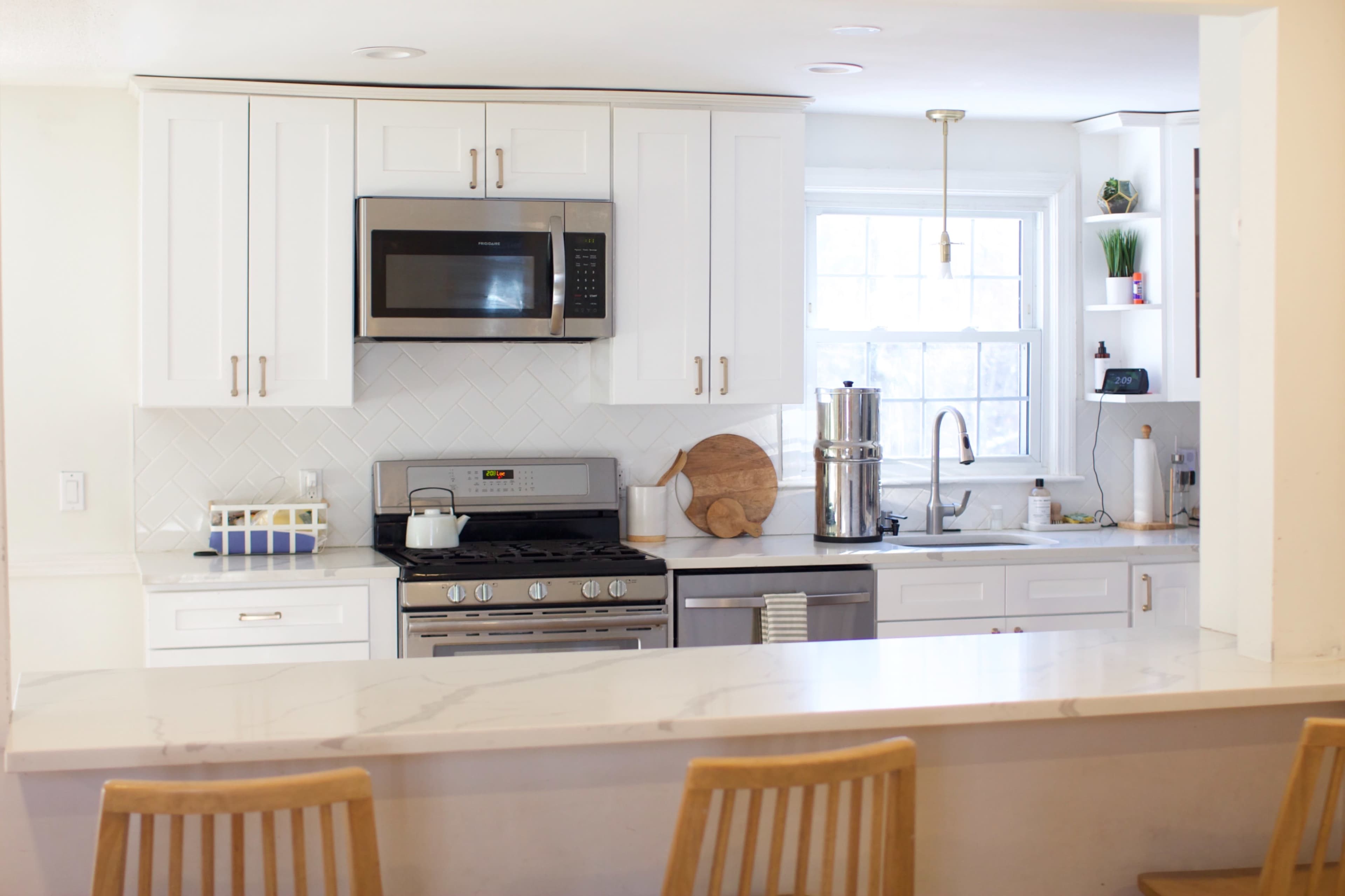 The image shows a modern kitchen with white cabinets, stainless steel appliances, and a countertop with bar stools.