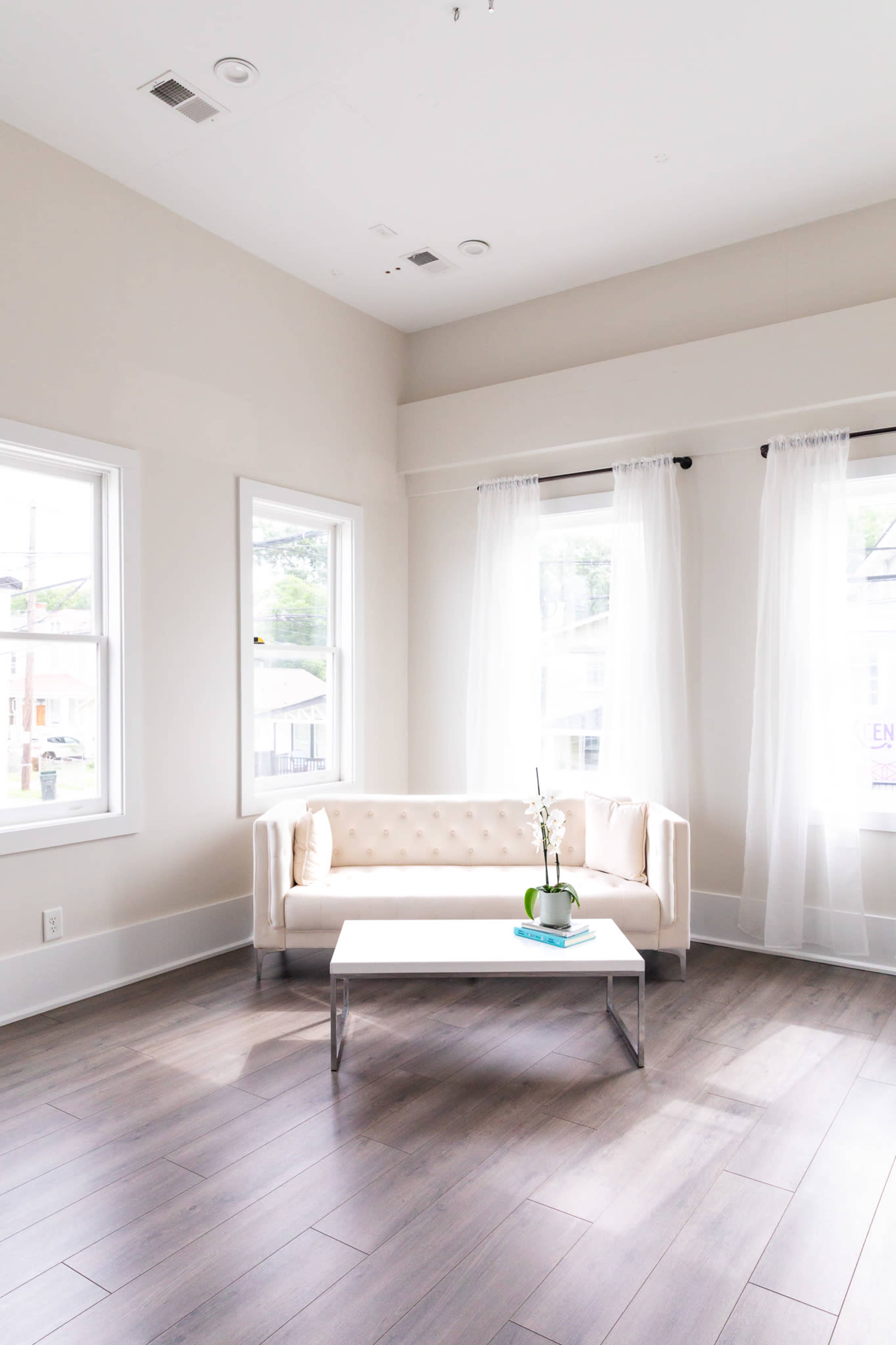 A light-filled living room features a beige tufted sofa, a white coffee table, and large windows with sheer curtains.