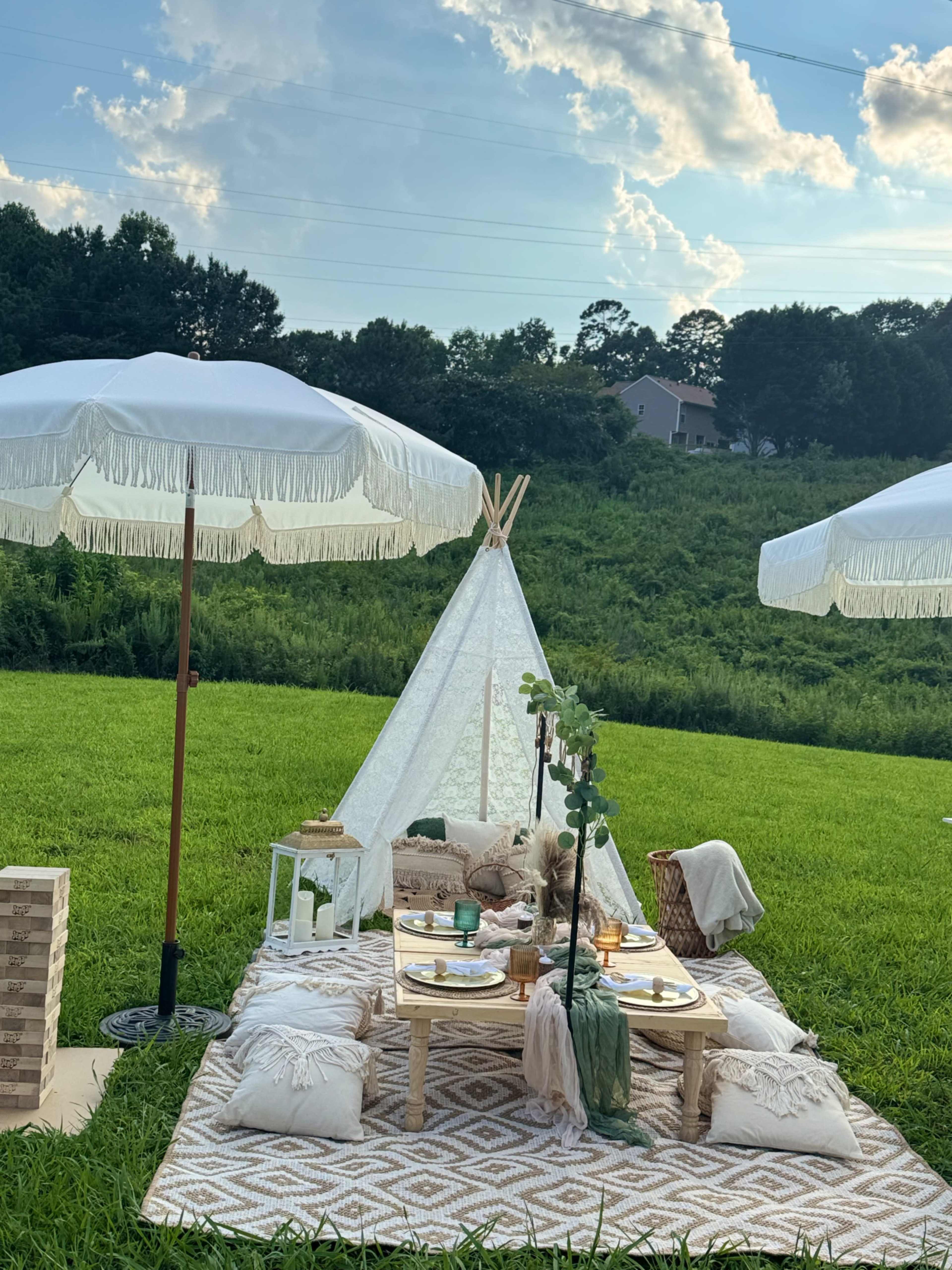 The scene features a cozy outdoor picnic setup with a teepee, low tables, cushions, and large umbrellas on a grassy field.