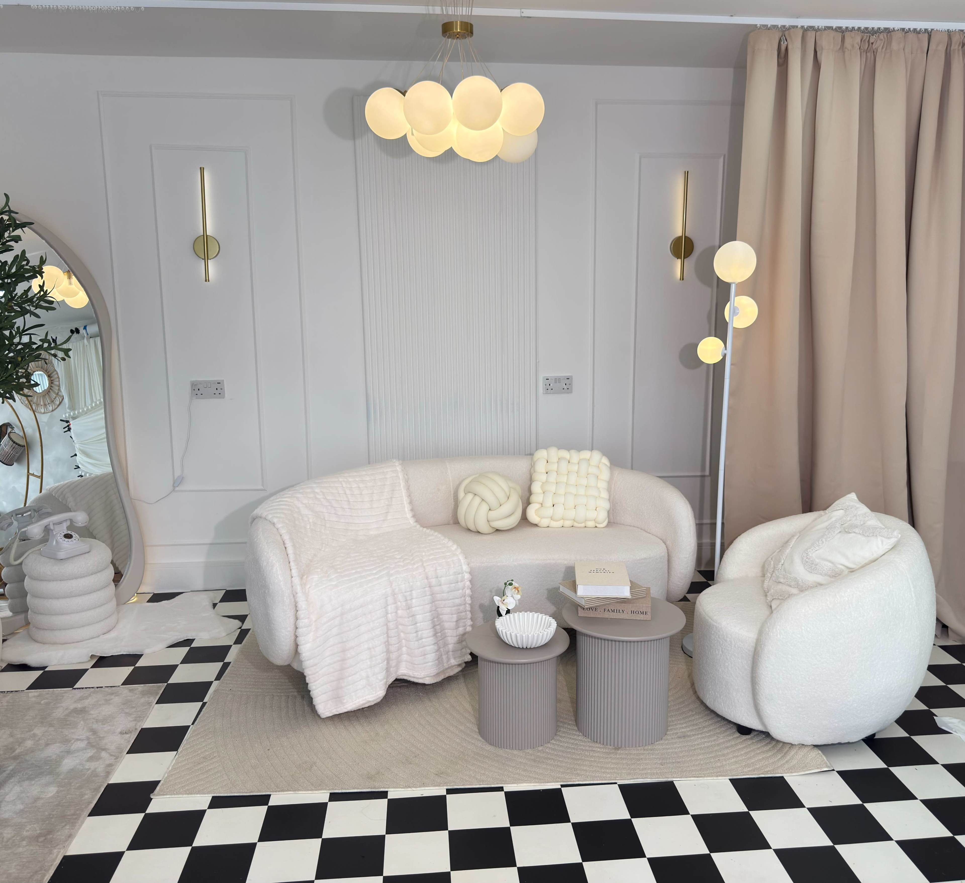 The image shows a cozy interior scene featuring a curved white upholstered sofa and a matching armchair, accompanied by two round, textured side tables on a black and white checkered floor.