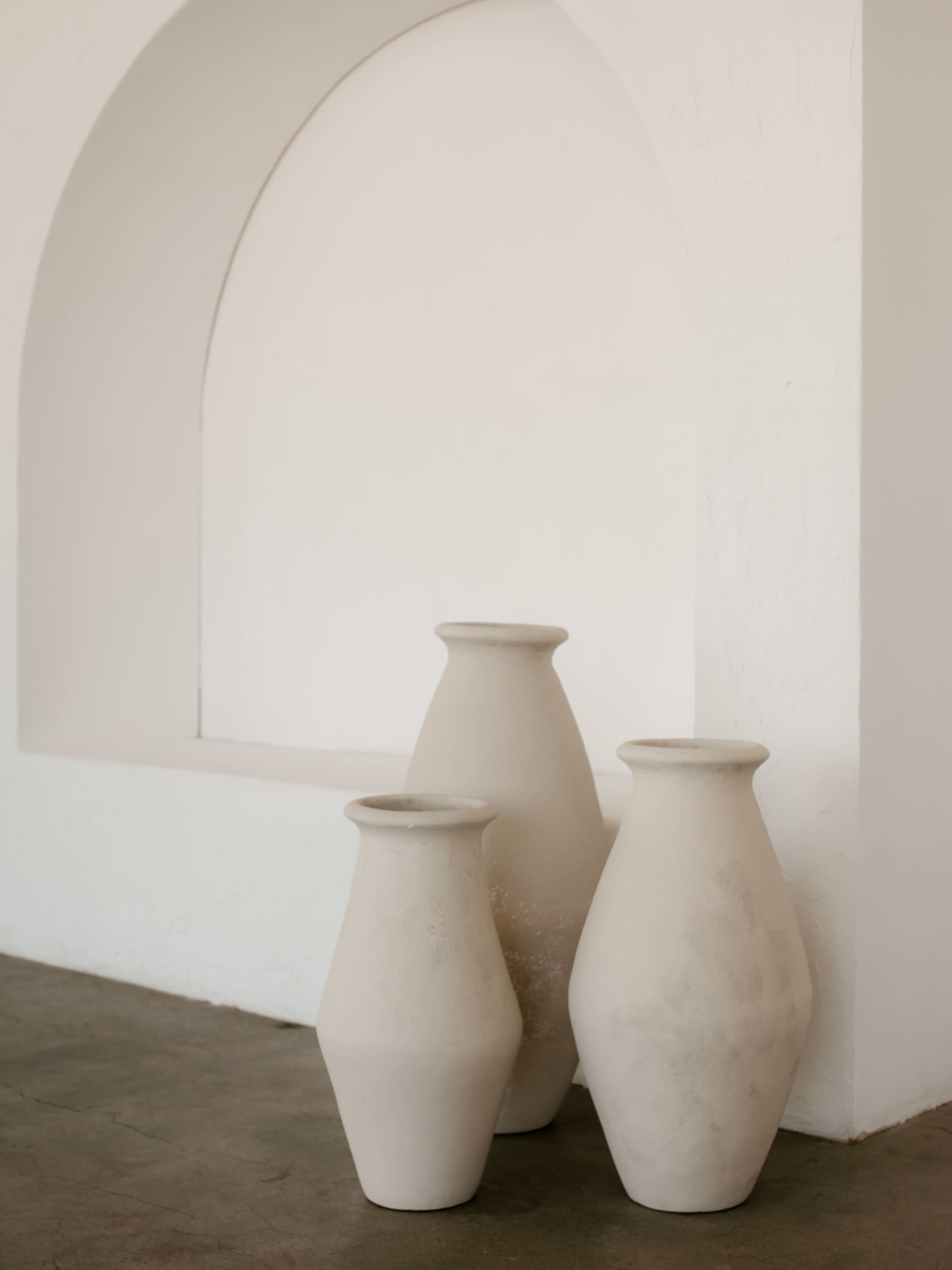 Three light-colored ceramic vases are arranged against a white wall with an arched niche.