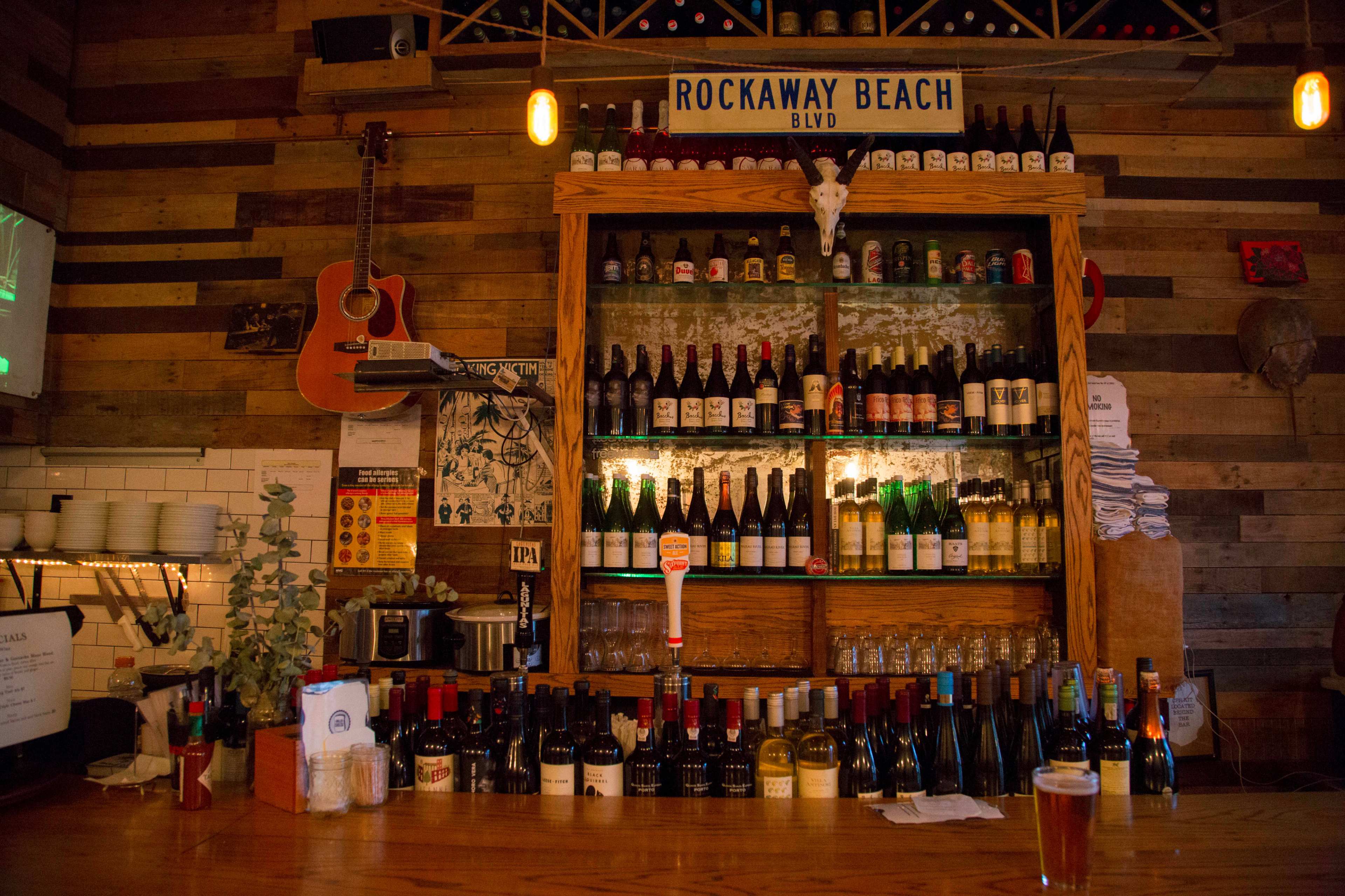 A wooden bar with an assortment of bottled beverages arranged on shelves behind it, accompanied by a guitar hanging on the wall.