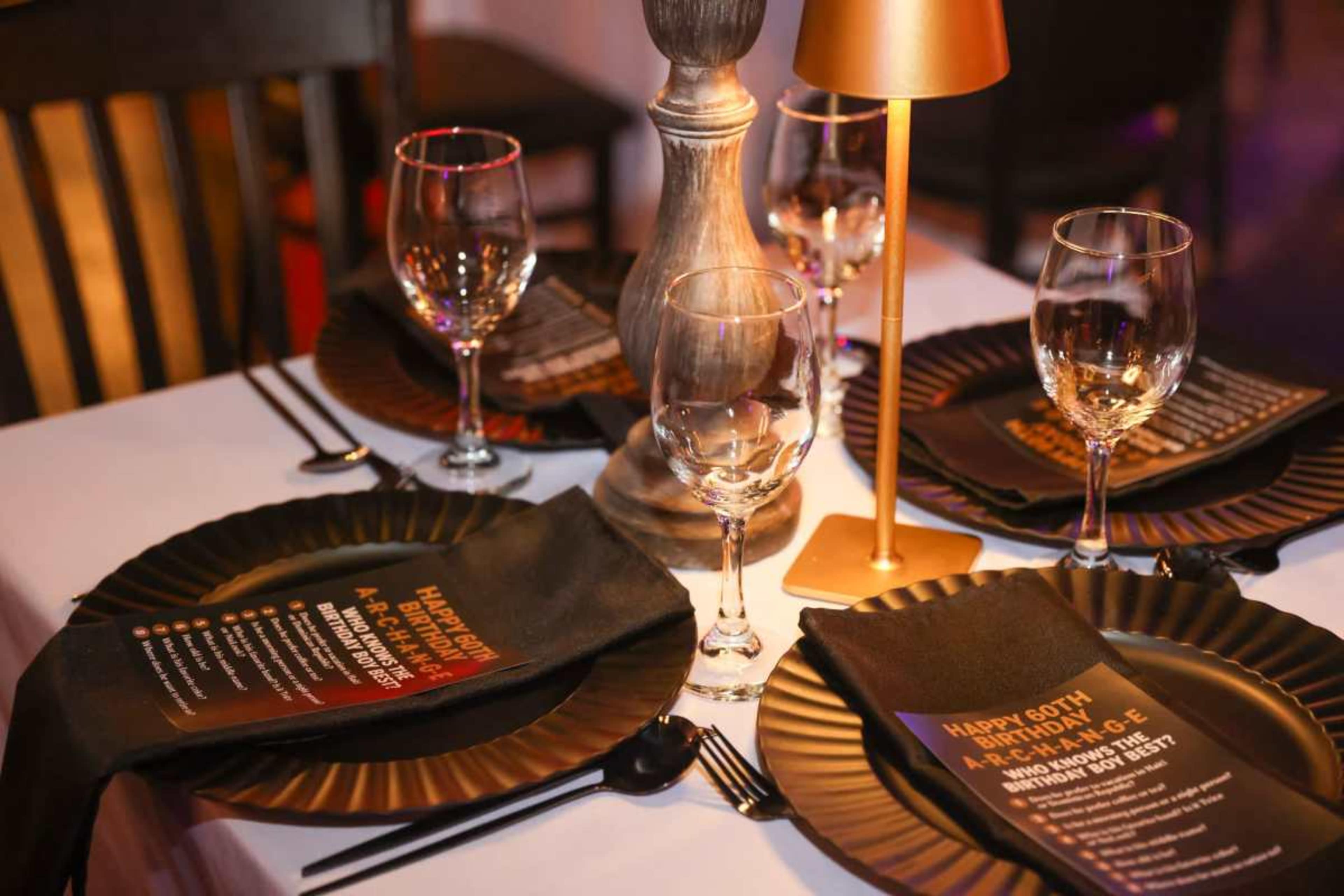 The table is set with elegant black plates, wine glasses, and folded napkins, featuring a birthday celebration theme with a golden candle holder as a centerpiece.