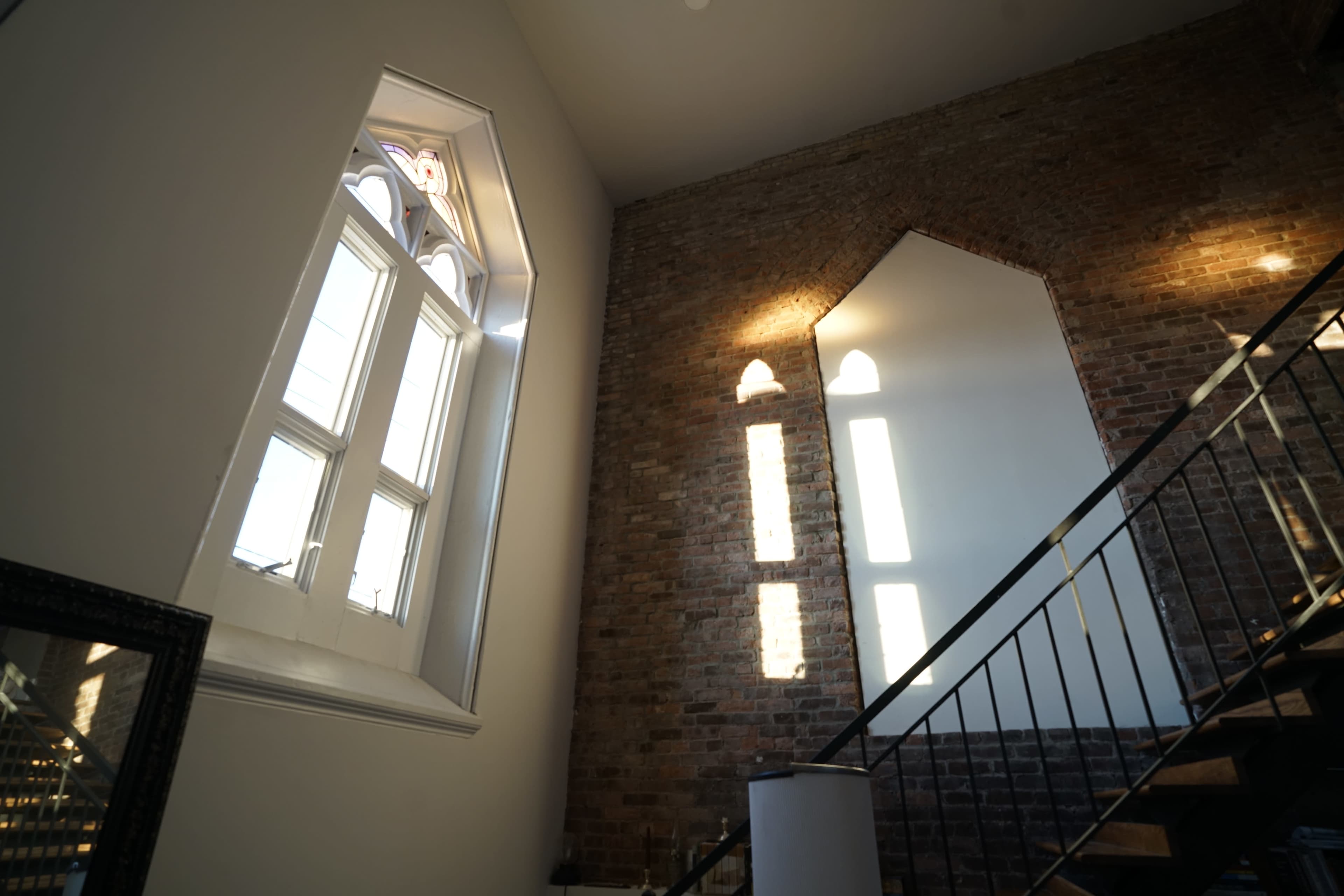 A stairway in a room with a brick wall and large arched windows casting shadows on the white walls.