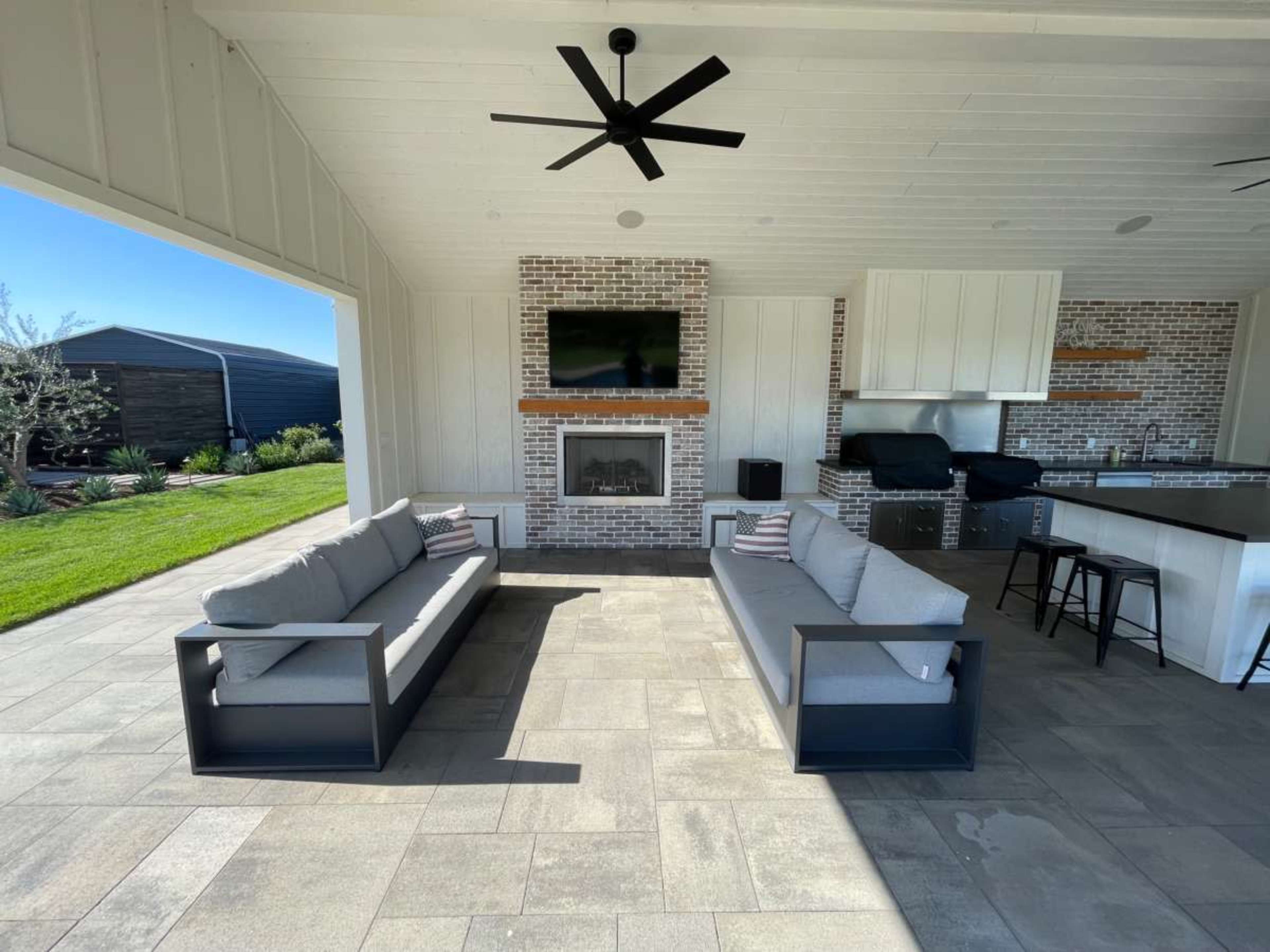 The image shows a covered outdoor seating area with two modern sofas facing each other, a brick fireplace, and a grill station in the background.