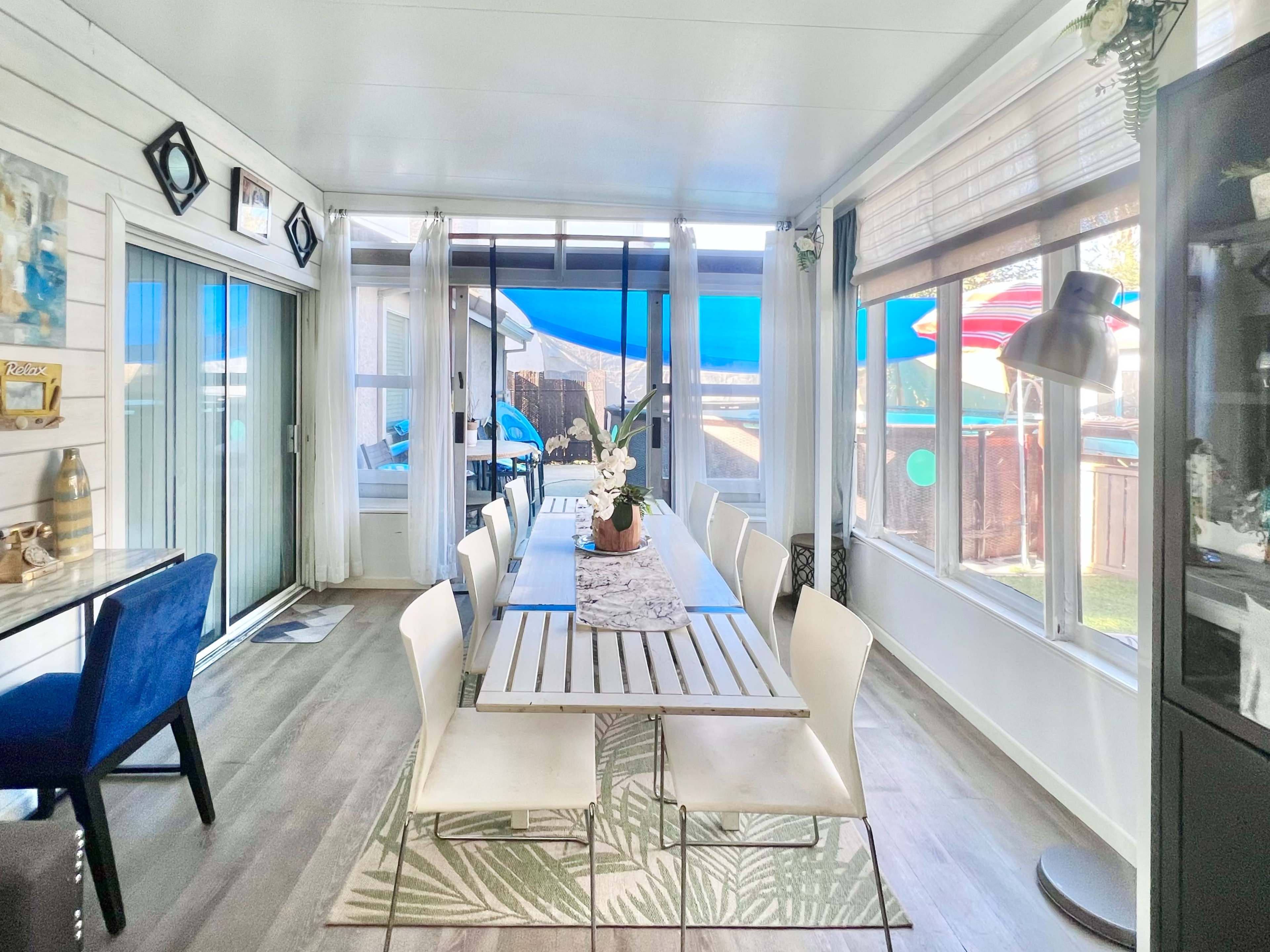 A bright sunroom featuring a long dining table with white chairs, surrounded by large windows and indoor plants.
