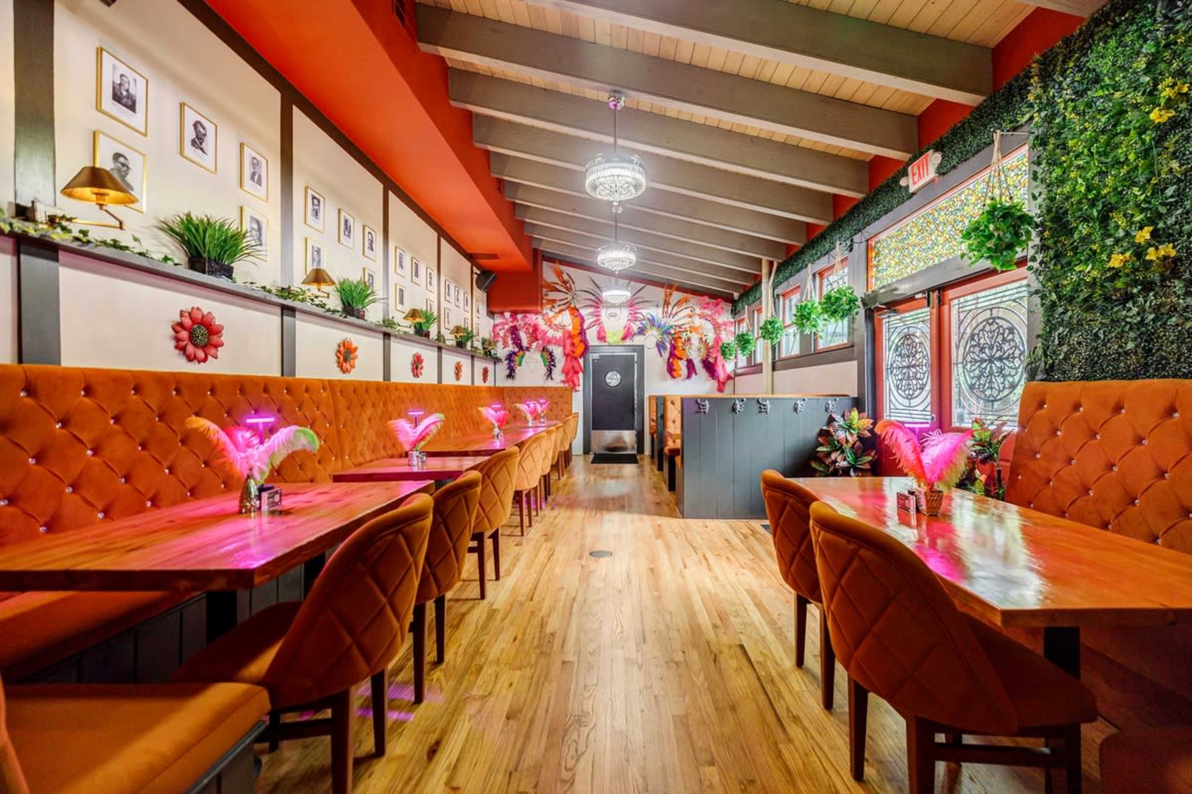 The interior of a restaurant featuring wooden tables, orange upholstered chairs, and decorative plants along the walls, with colorful lighting and framed photographs.