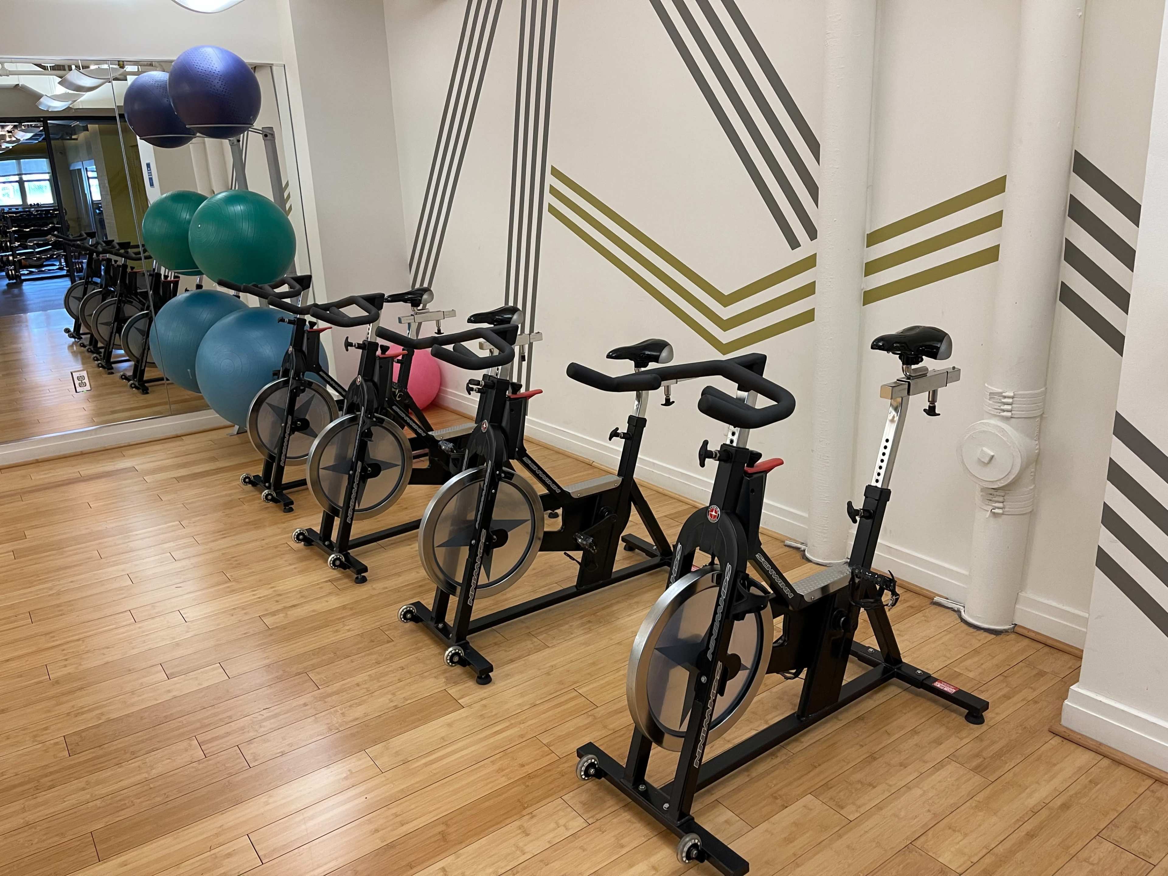 A row of stationary bikes lined up against a wall with exercise balls and a large mirror in a fitness studio.
