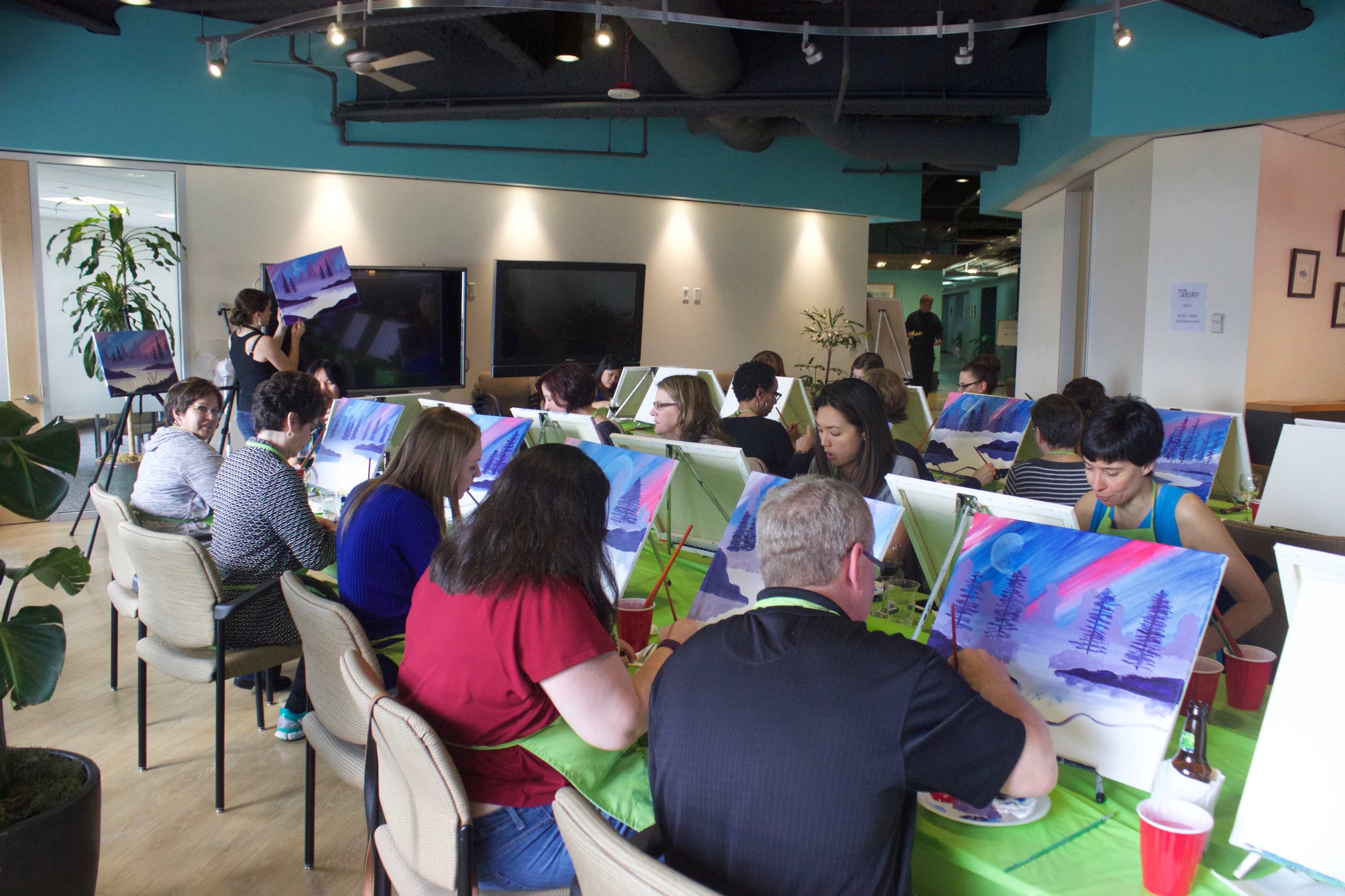 A group of people participates in a painting class, each working on a canvas while seated at tables in a well-lit studio.