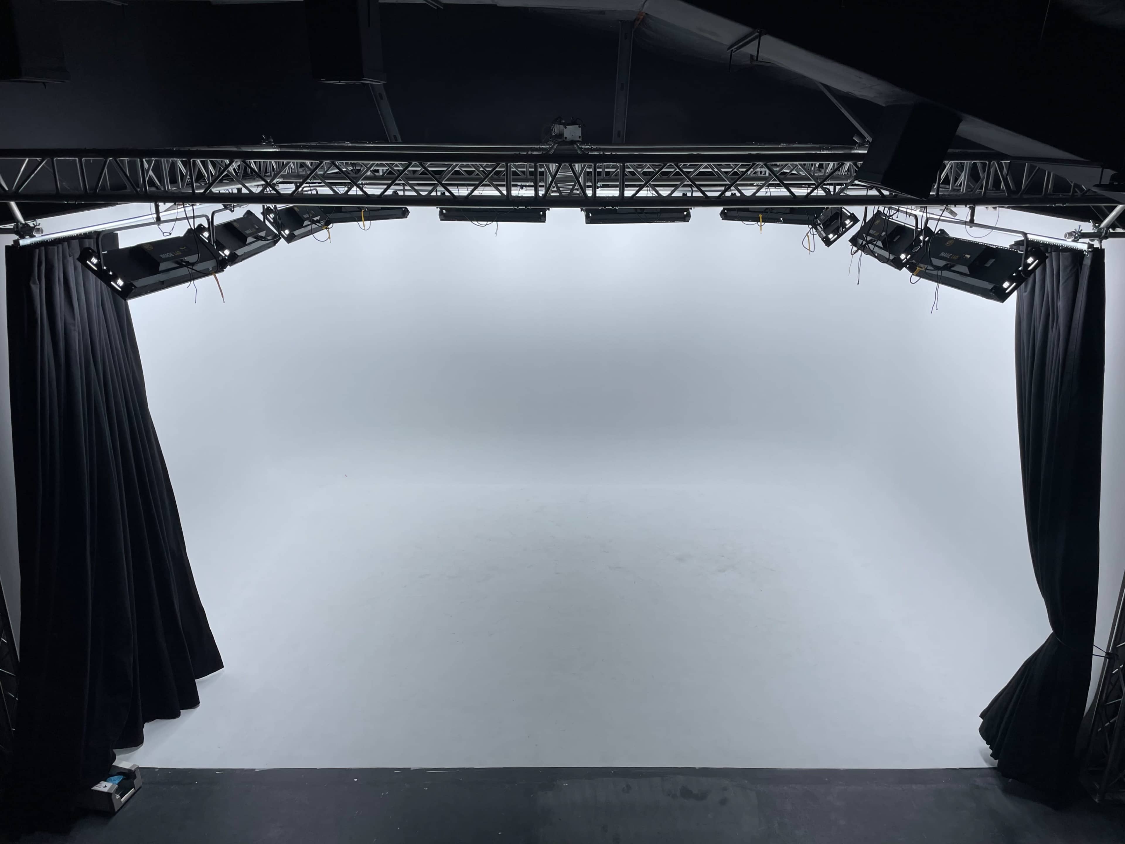 An empty stage with a white backdrop and black curtains on either side, illuminated by overhead lights.