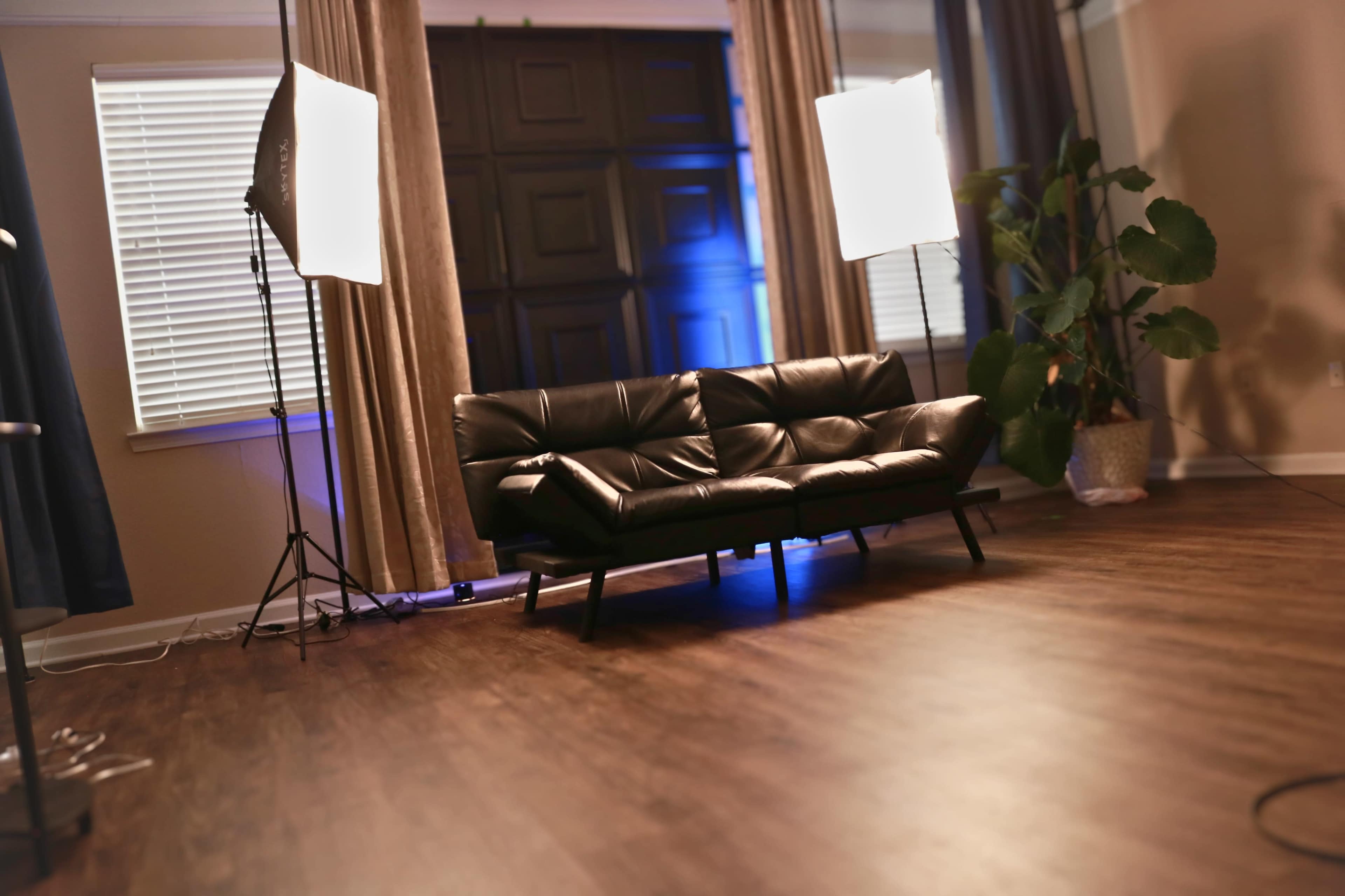 The image shows a modern black leather sofa positioned in a room with large windows, curtains, and studio lighting set up in the background.