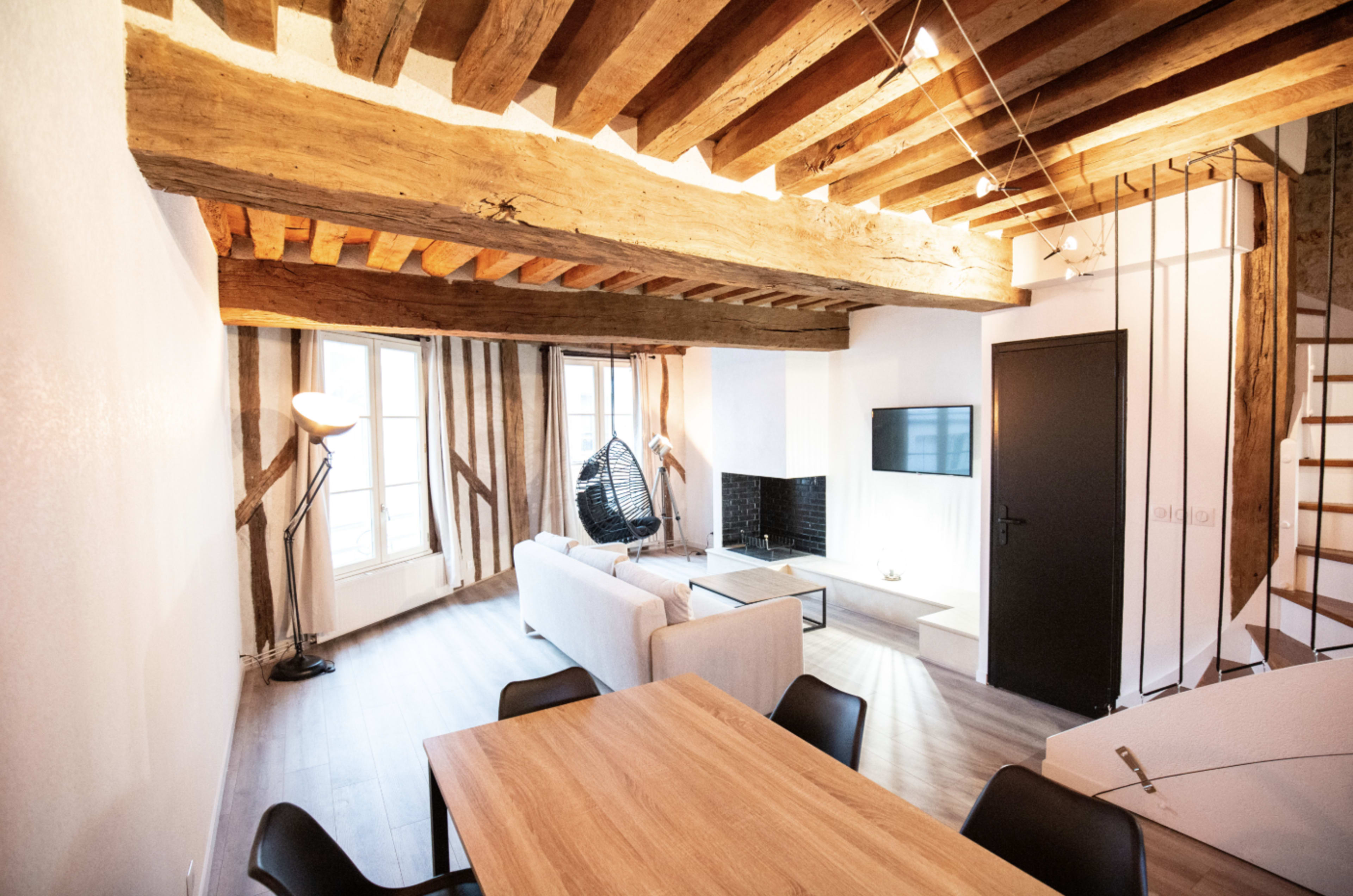 The image shows a modern living space featuring exposed wooden beams, a sofa, a dining table, and a staircase, with large windows allowing natural light.