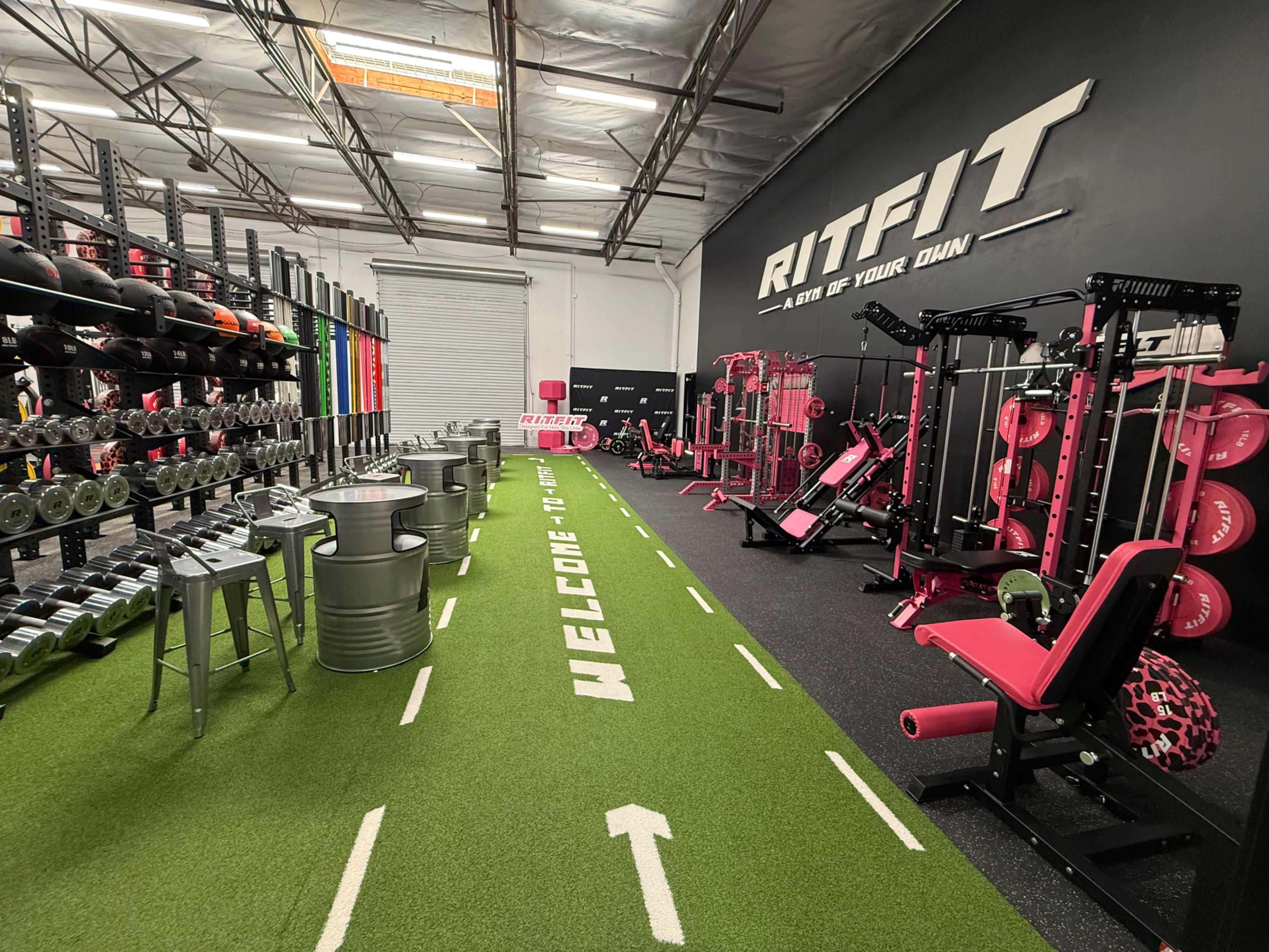 The image shows a fitness gym featuring various exercise equipment, weights, and a turf pathway with a welcome message.
