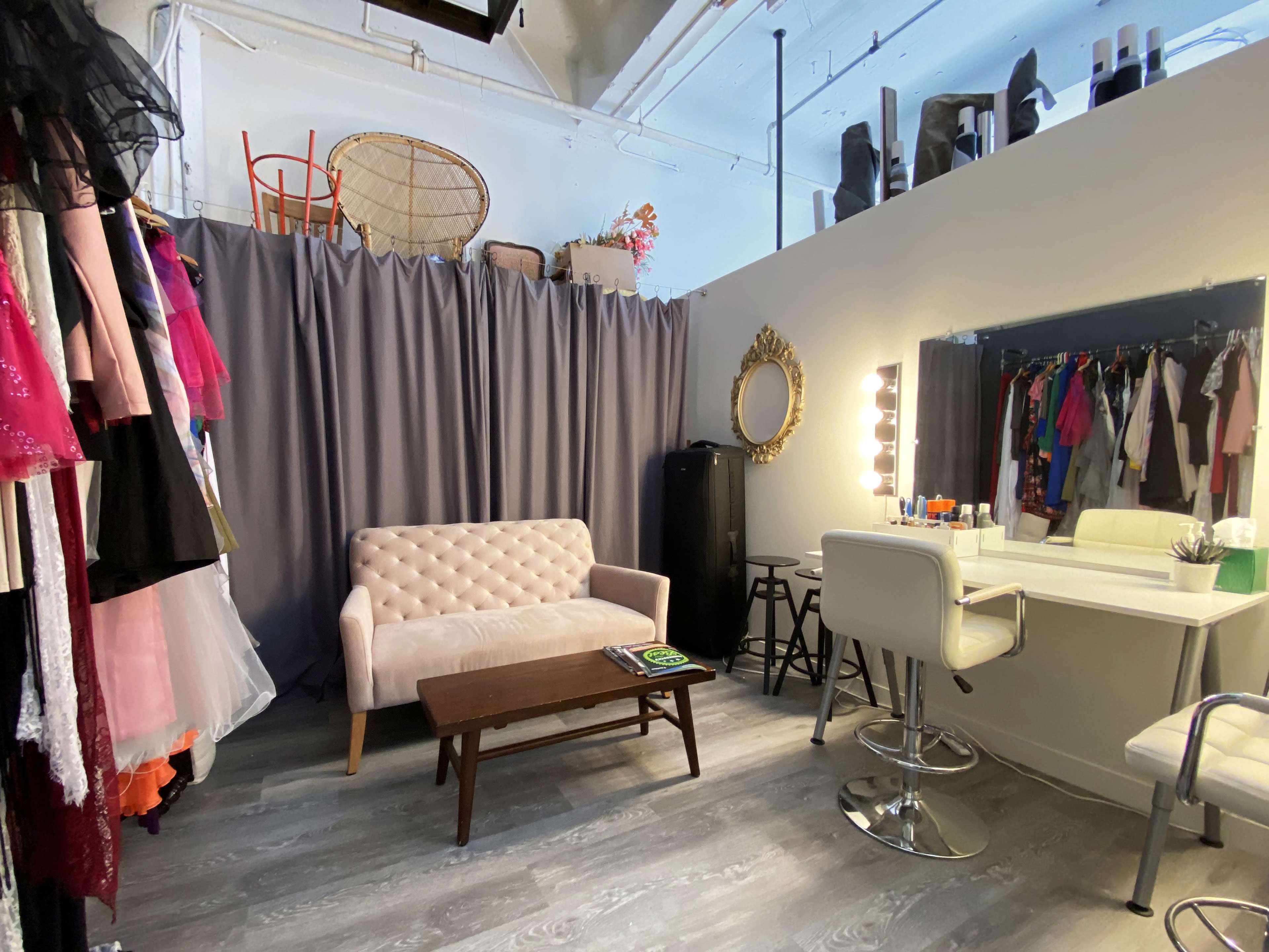The image shows a dressing room with a pink sofa, a wooden coffee table, a vanity with bright lights, and clothing hanging on the wall behind a gray curtain.
