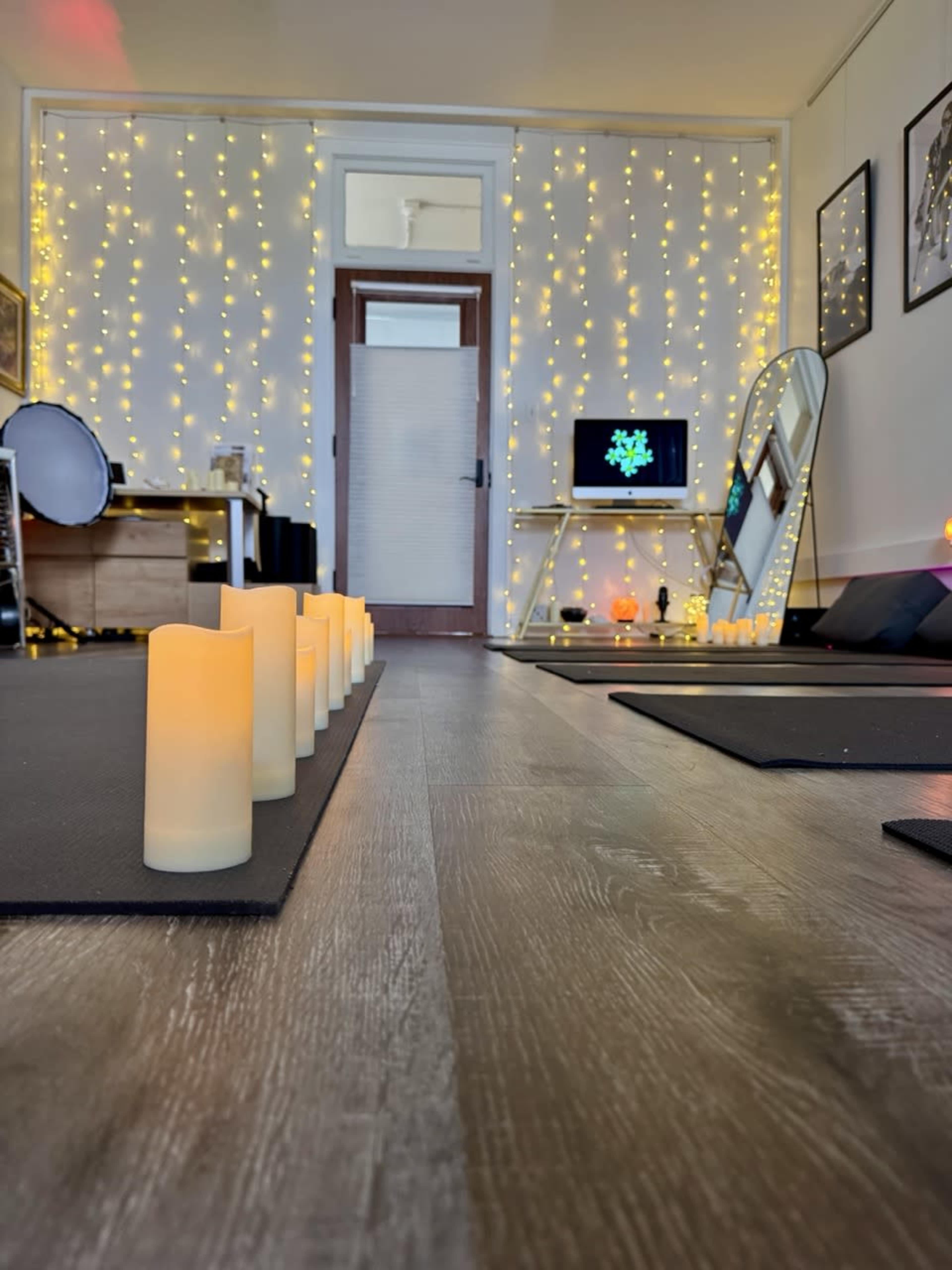The image shows a yoga studio with lit candles aligned on a black mat, twinkling fairy lights on the walls, and a mirror reflecting a digital screen.