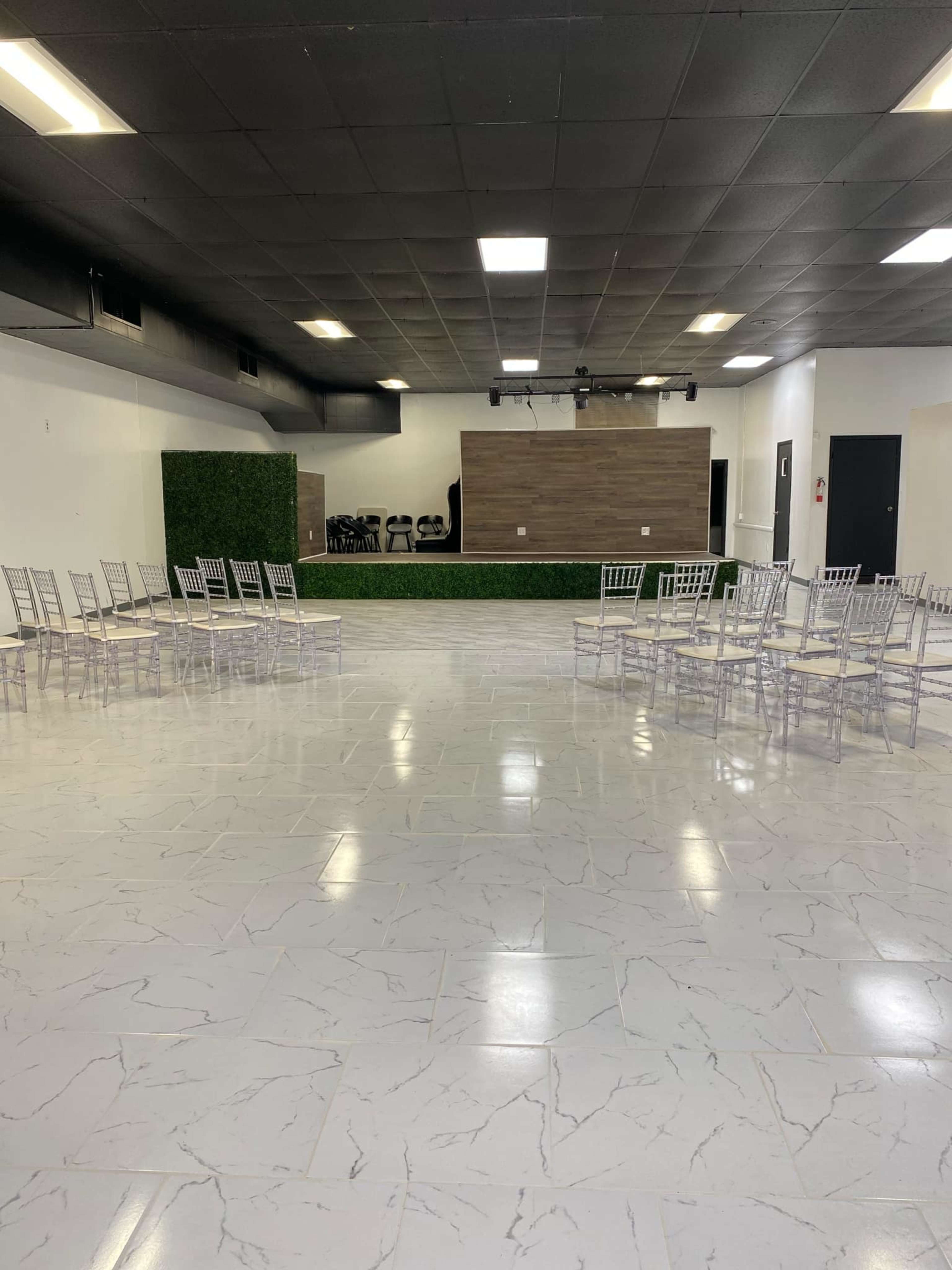 A spacious room with white tiled flooring, a stage at the front, and two rows of clear chairs arranged facing the stage.