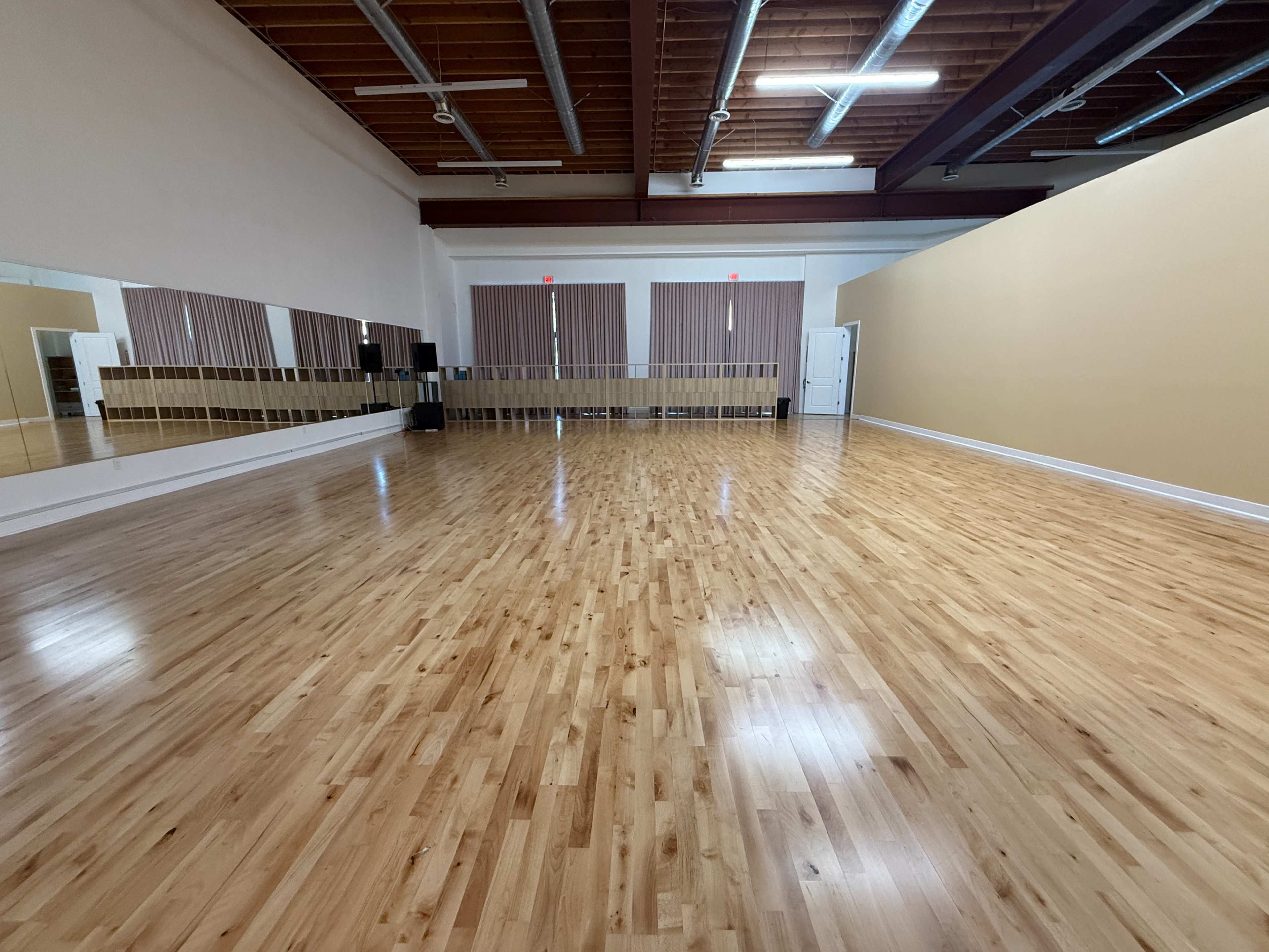 The image shows an empty dance studio with wooden flooring, large mirrors on one wall, and natural light coming from overhead lights.