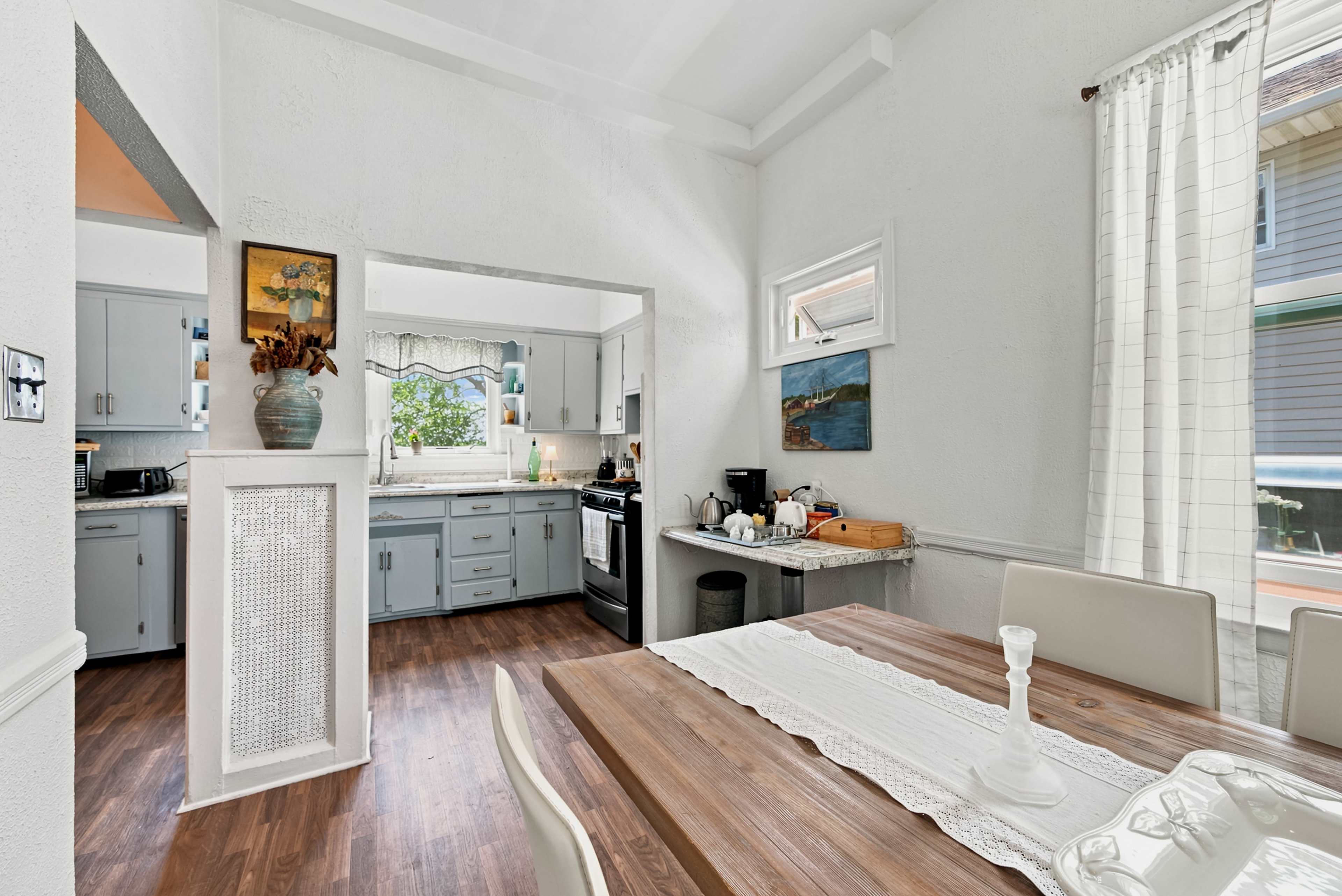 The image shows a bright kitchen and dining area with light-colored walls, modern cabinetry, and a wooden dining table set for a meal.