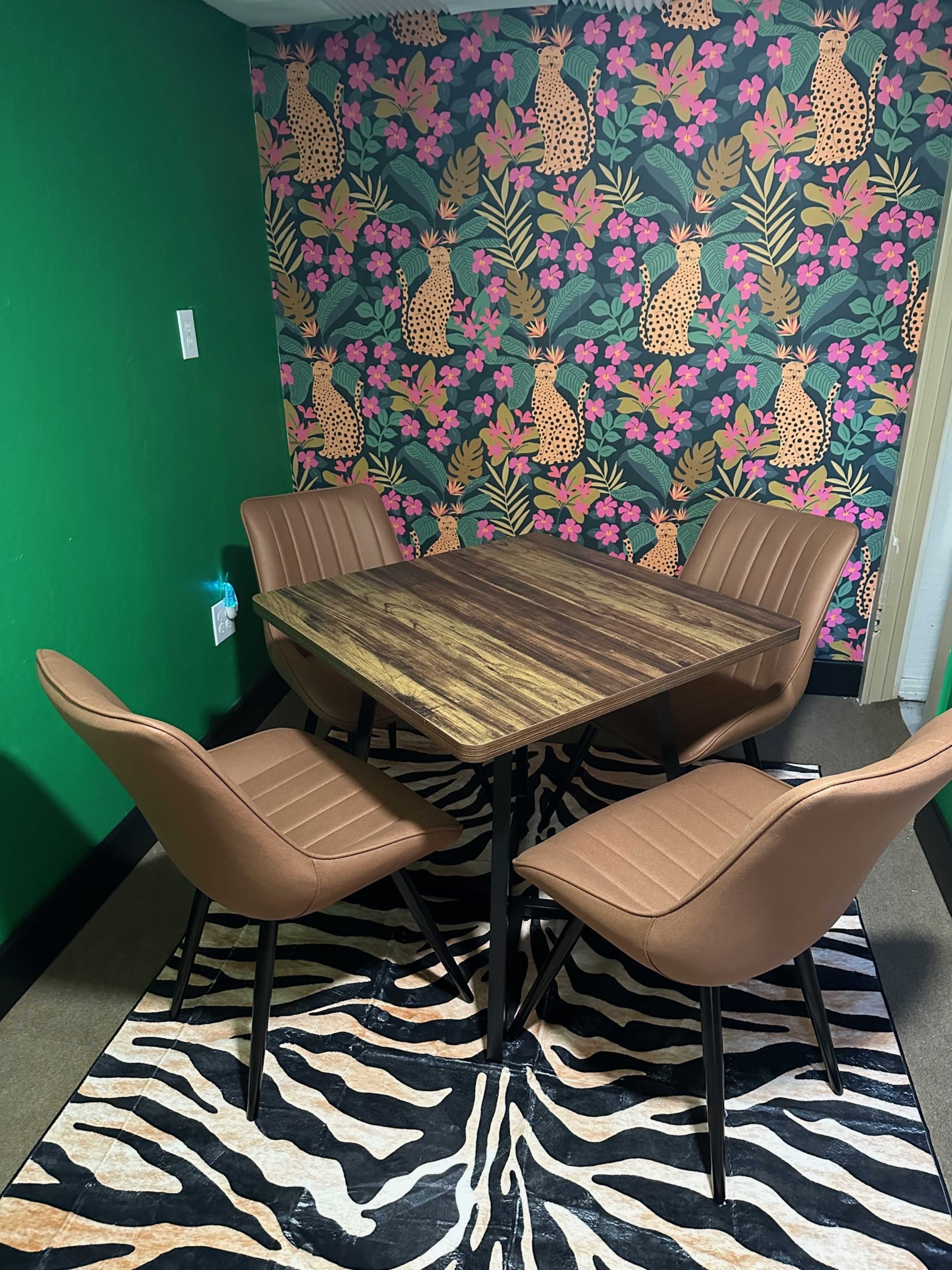 The image shows a small dining area with a wooden table surrounded by four brown chairs, set against a vibrant floral and leopard-patterned wallpaper and a green wall.