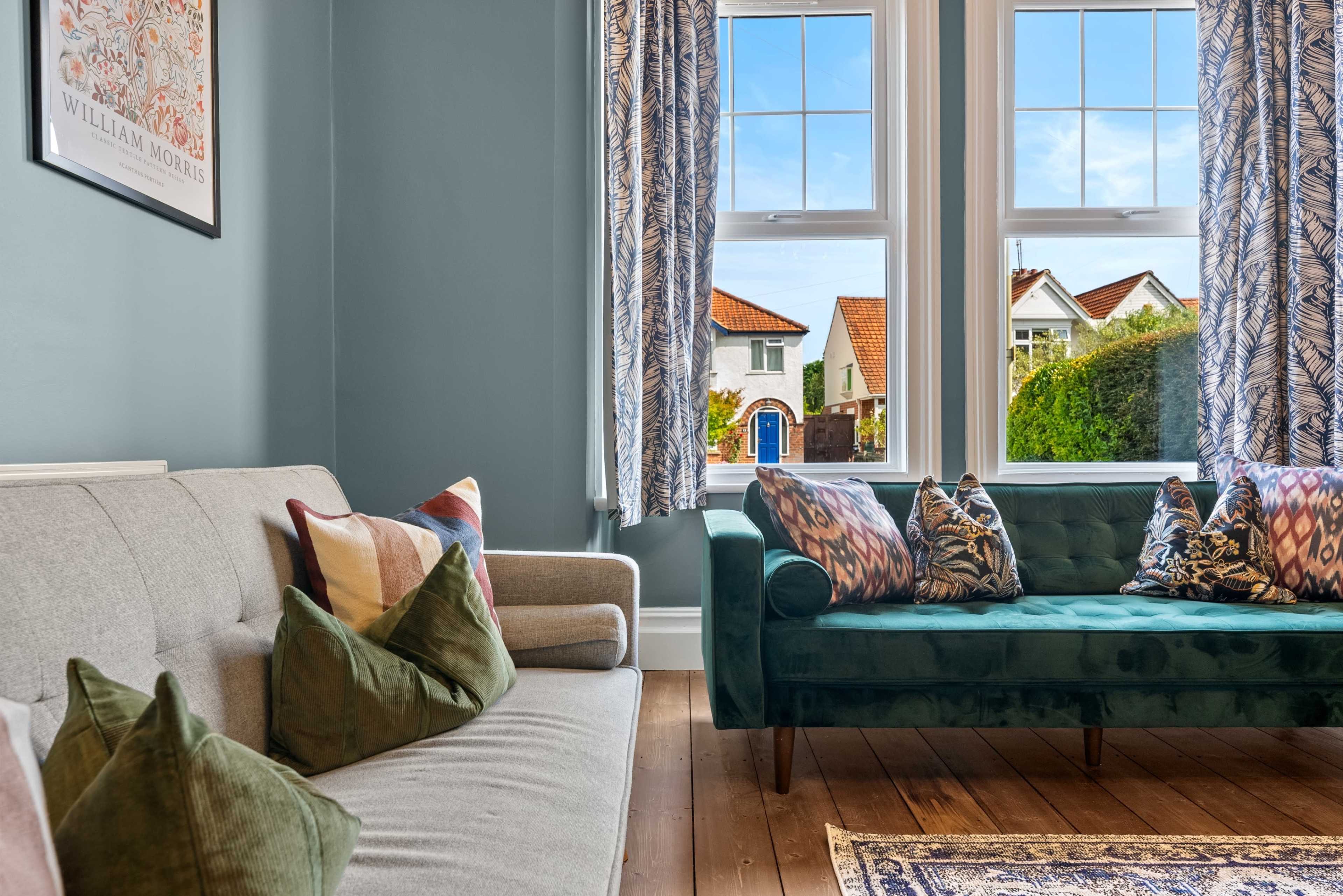A living room features a light gray sofa and a dark green couch with decorative pillows, set against blue walls and large windows that show a view of a house and garden outside.