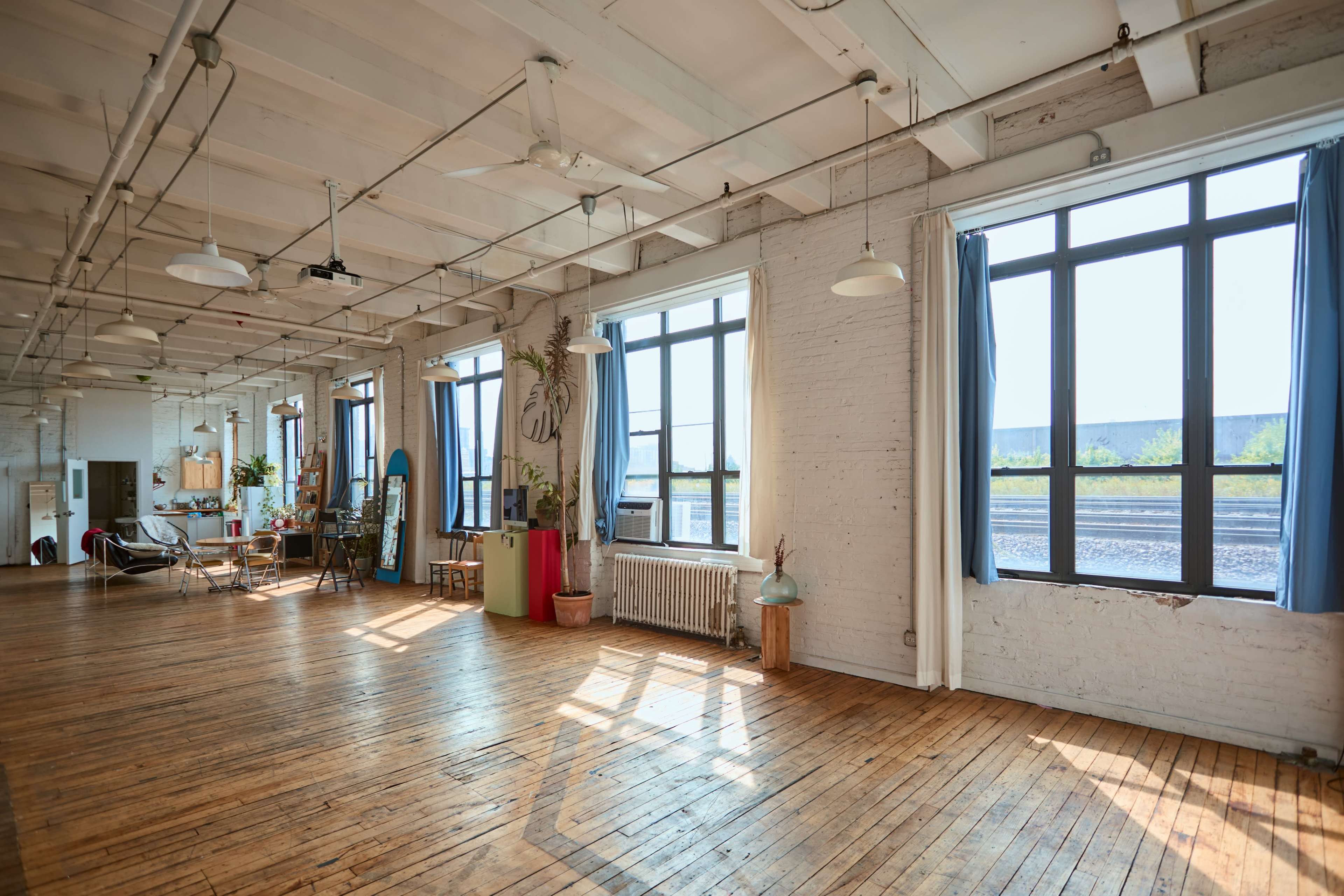 The image shows a spacious, well-lit interior with large windows, wooden floors, and various furniture arranged along the walls.