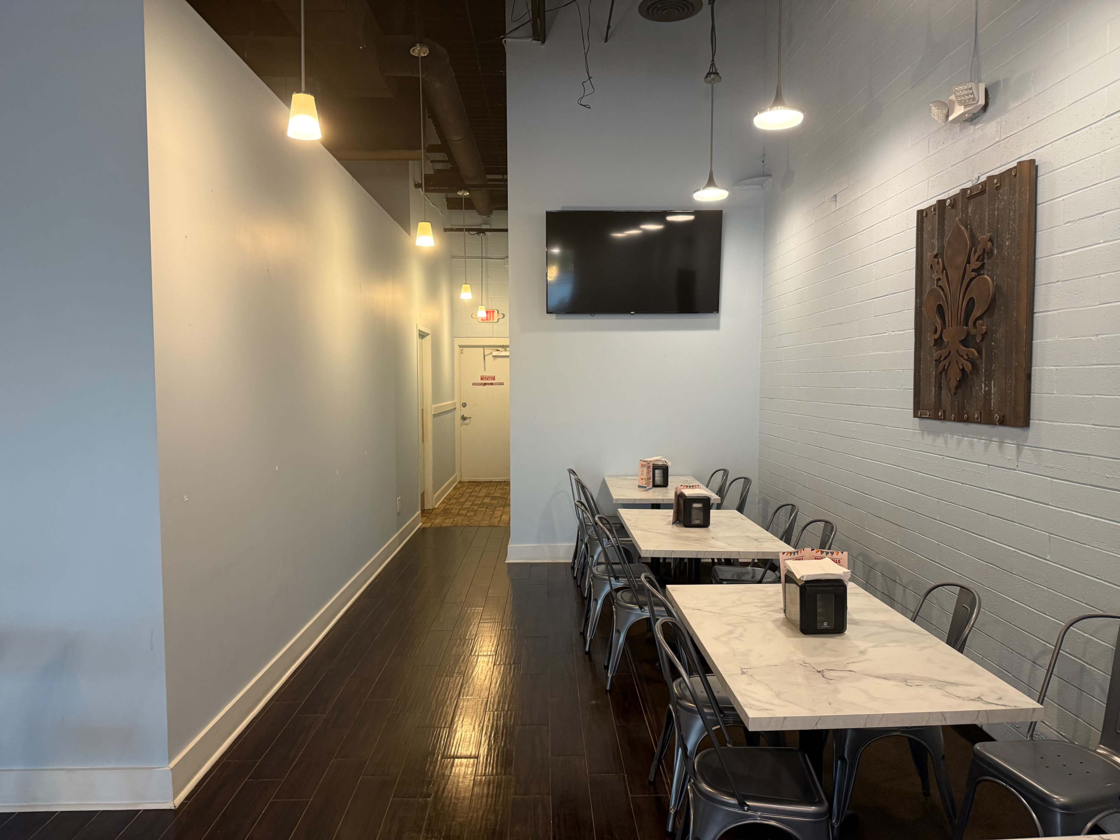 The image shows a narrow dining area with a long marble table lined with metal chairs, illuminated by pendant lights and featuring a wall-mounted television.