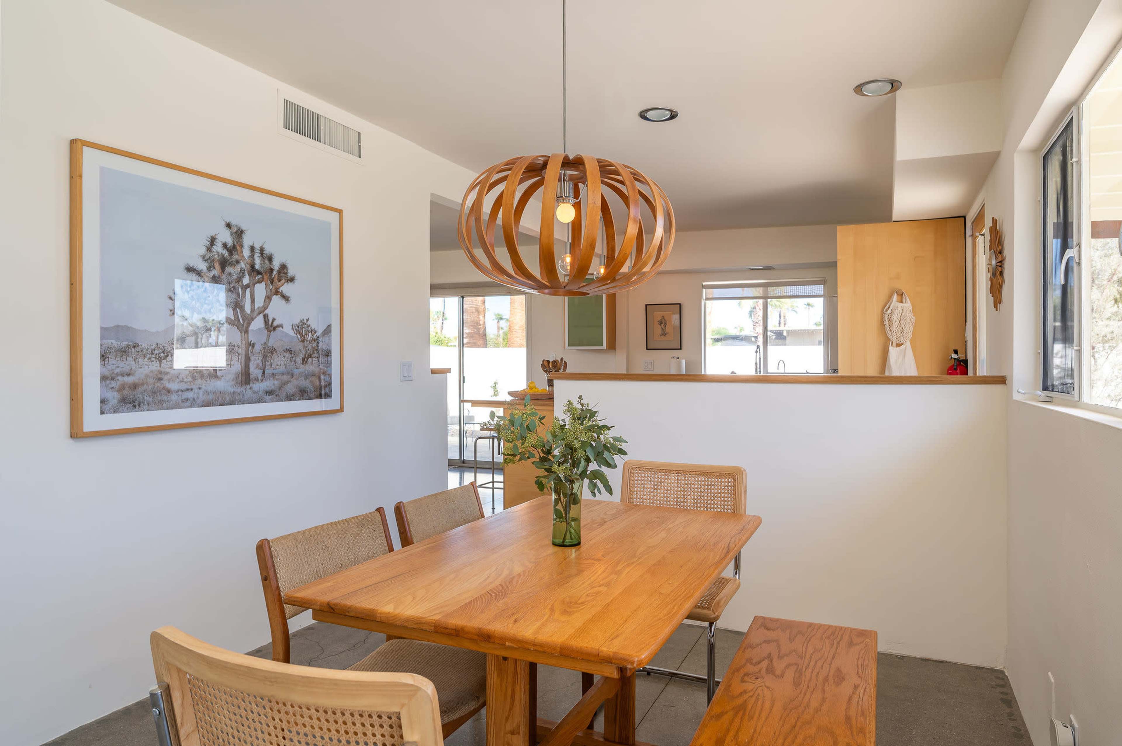 A dining area features a wooden table surrounded by chairs and a pendant light, with a framed landscape photo of a Joshua tree on the wall.