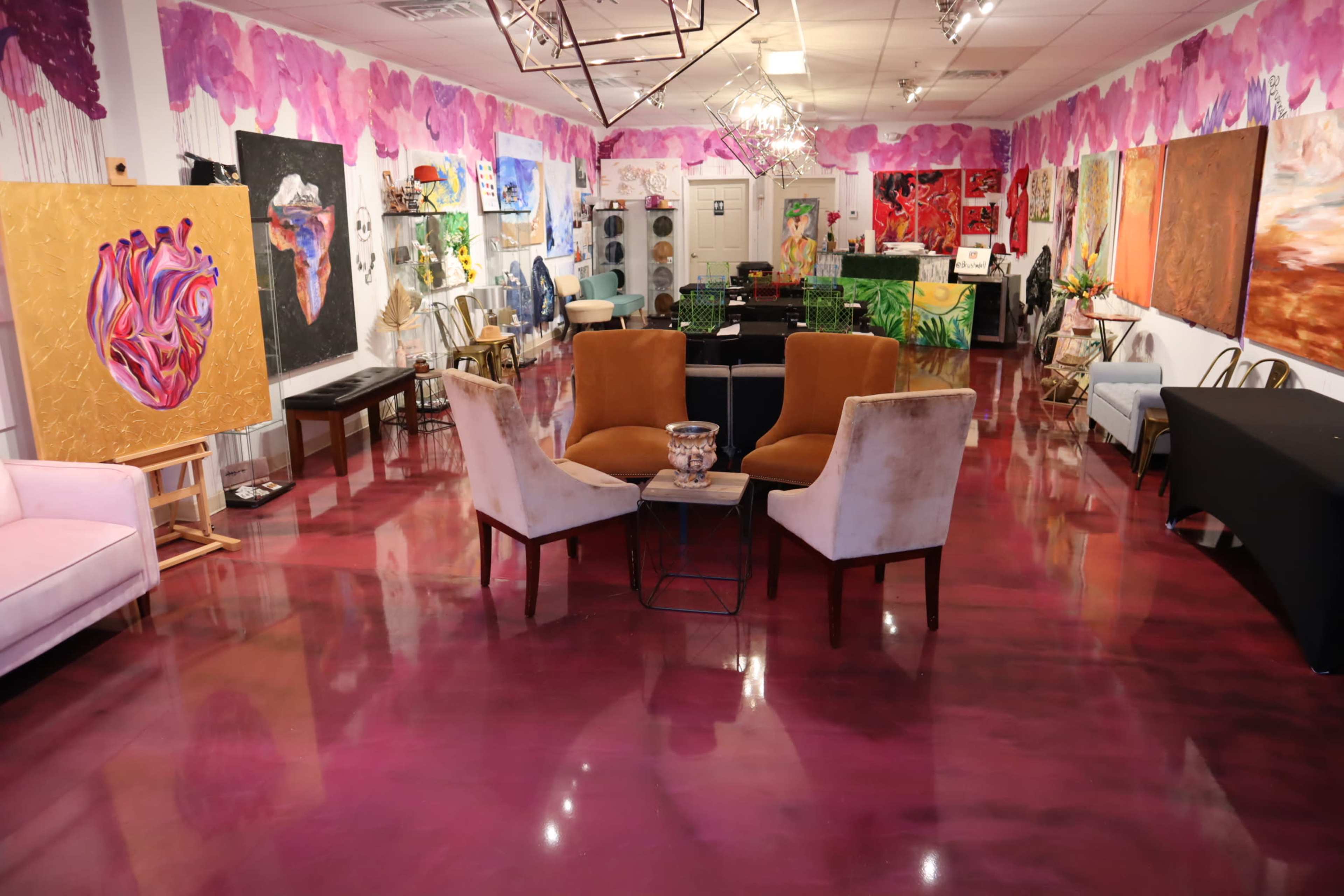 An art gallery filled with colorful paintings on the walls, two brown chairs and a small table in the center, and various seating arrangements throughout.