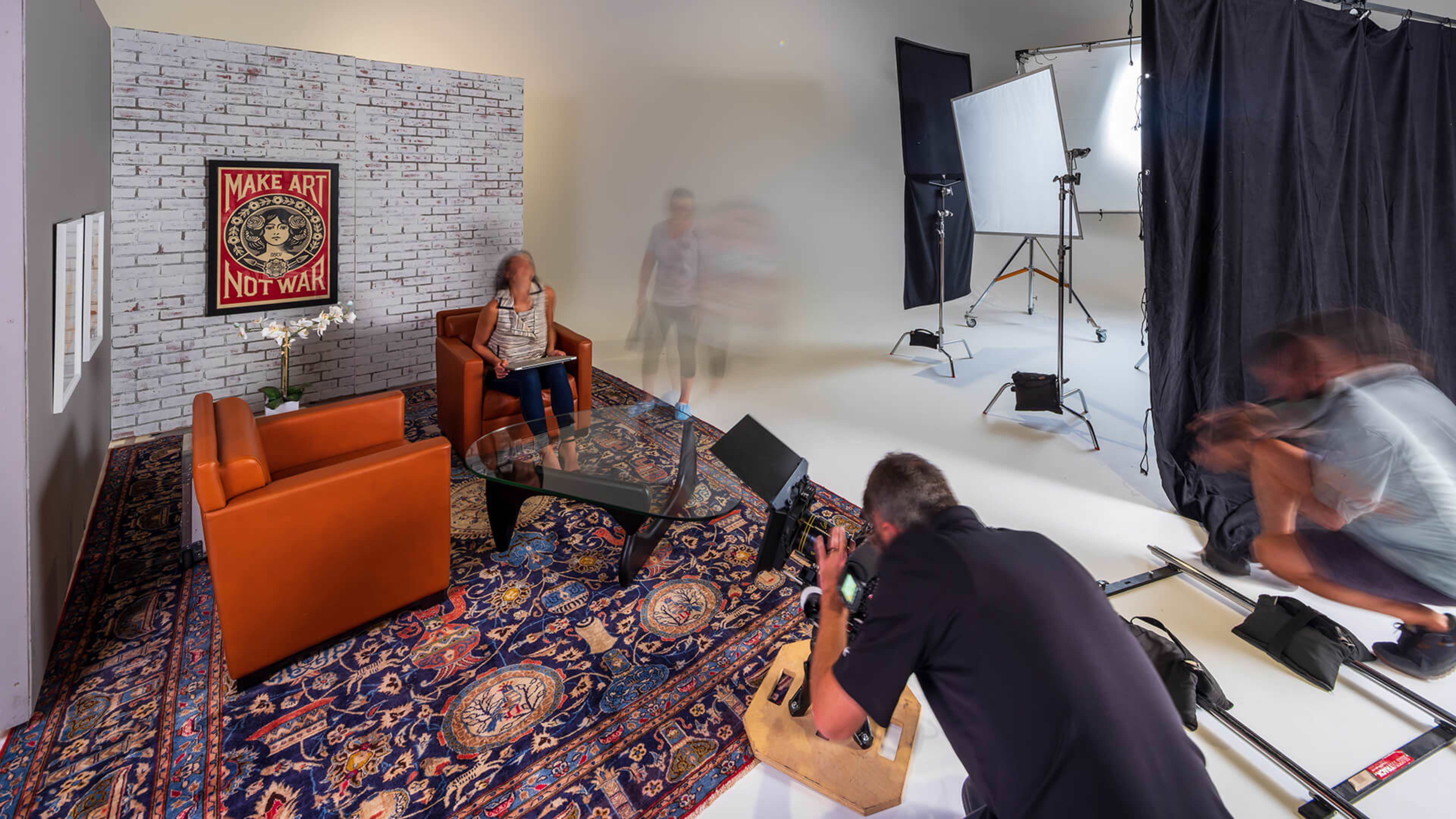 A photography studio features a woman sitting on a couch with a decorative rug, while a crew sets up equipment in the background.